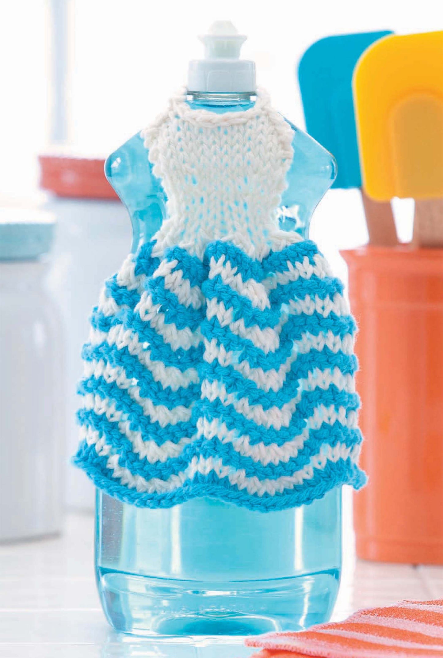 The Leisure Arts "Dishcloth Dresses—The Cutest Quick & Easy Gifts, Ever!" digital download shows a blue and white zigzag knit bottle cover styled like a doll outfit, with kitchen utensils and containers blurred in the background.