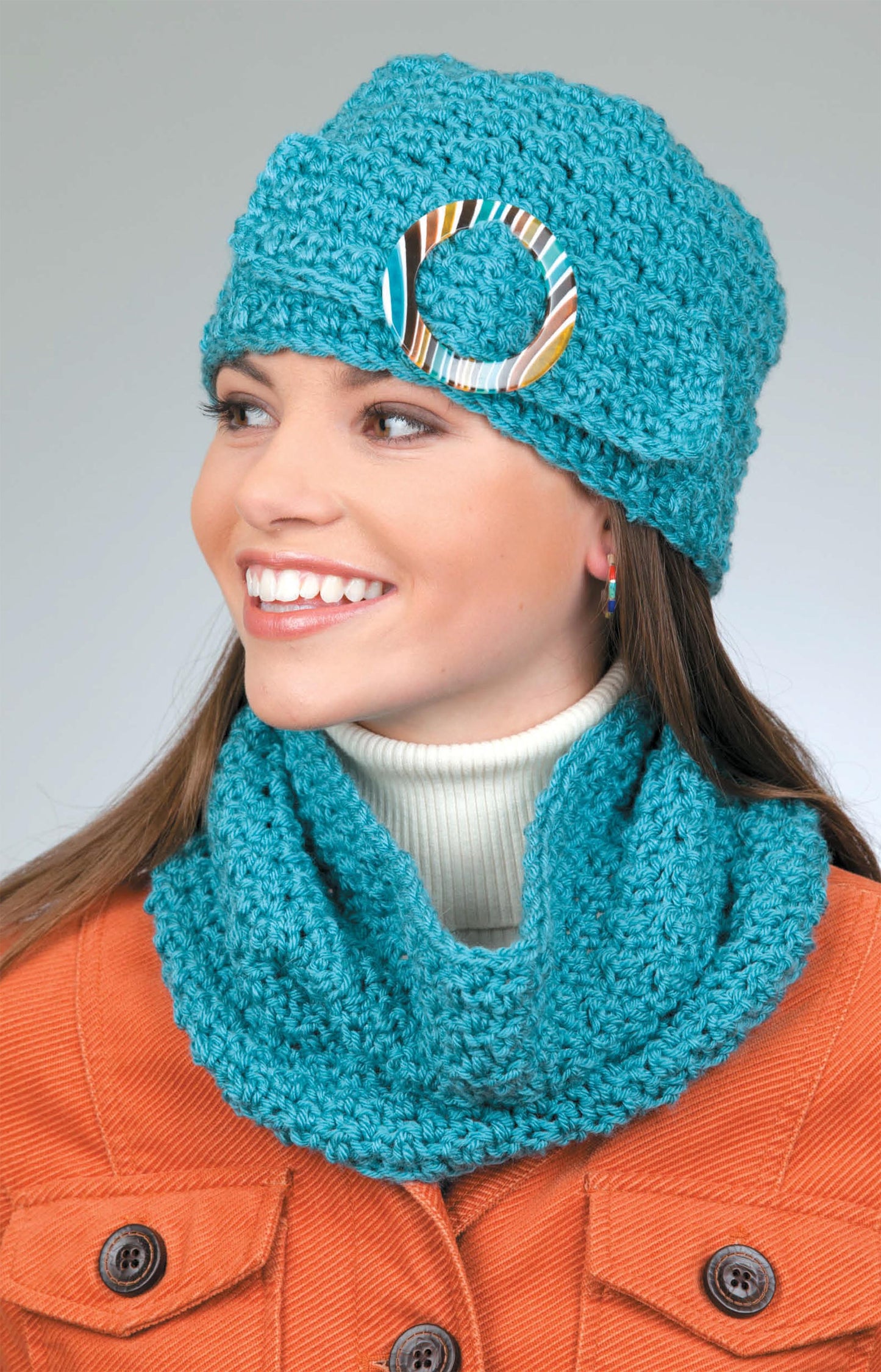 A woman wears Leisure Arts' Hats & Scarves set—a turquoise crochet hat with a large ring and matching cowl—paired with a white turtleneck and orange jacket, set against a plain background.