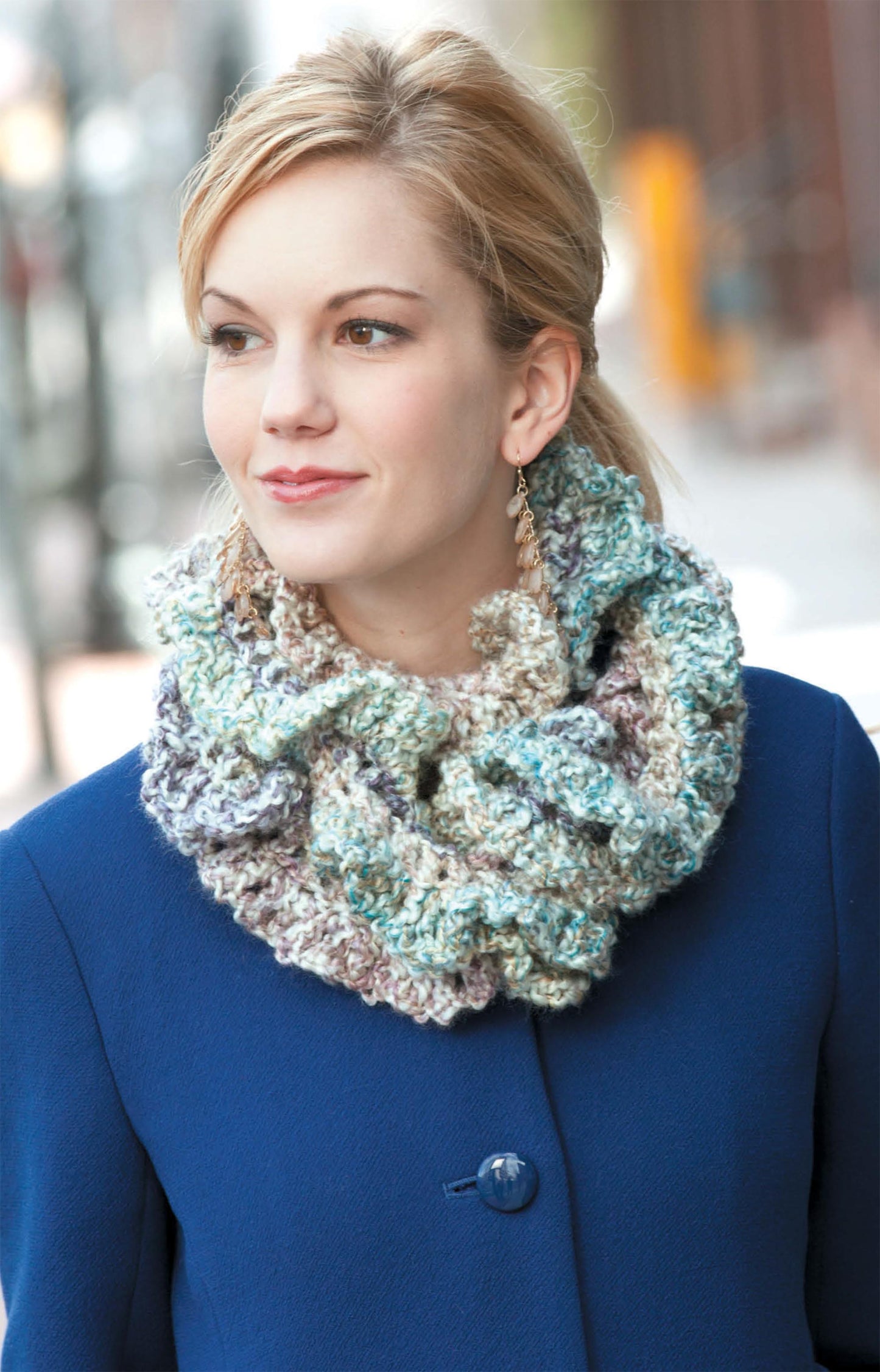 A woman in a blue coat and chunky crochet scarf stands outdoors, smiling. Her light hair is pulled back, and she wears dangling earrings. Explore cozy winter style with Hats & Scarves from Leisure Arts.