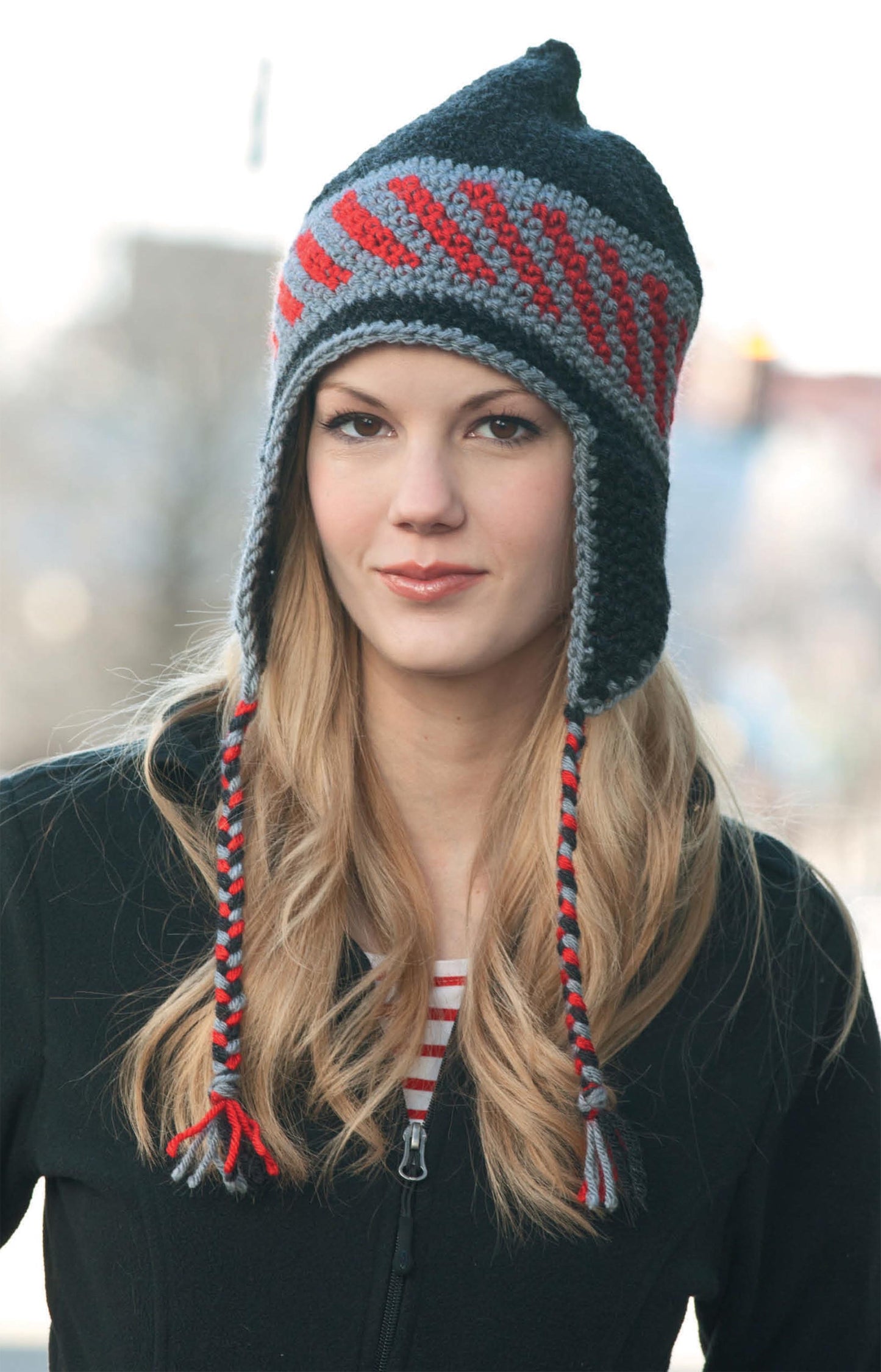 A woman with long blonde hair models Leisure Arts' Hats & Scarves—a black and red knit earflap hat with braided tassels and a chic crochet scarf. She stands outdoors in a black jacket, set against a softly blurred background.