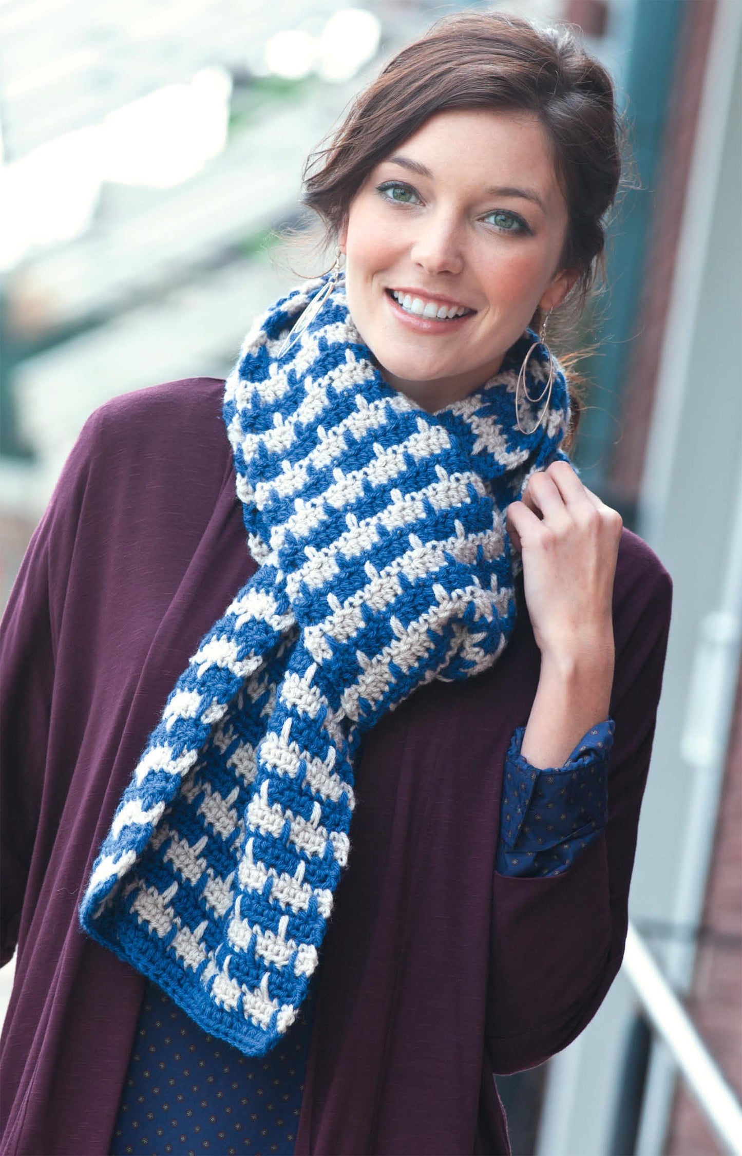 A woman smiles outdoors near a building, modeling a chunky blue and white geometric knit scarf from Leisure Arts' Hats & Scarves collection, holding the cozy accessory with one hand while wearing a purple jacket and blue polka-dot blouse.
