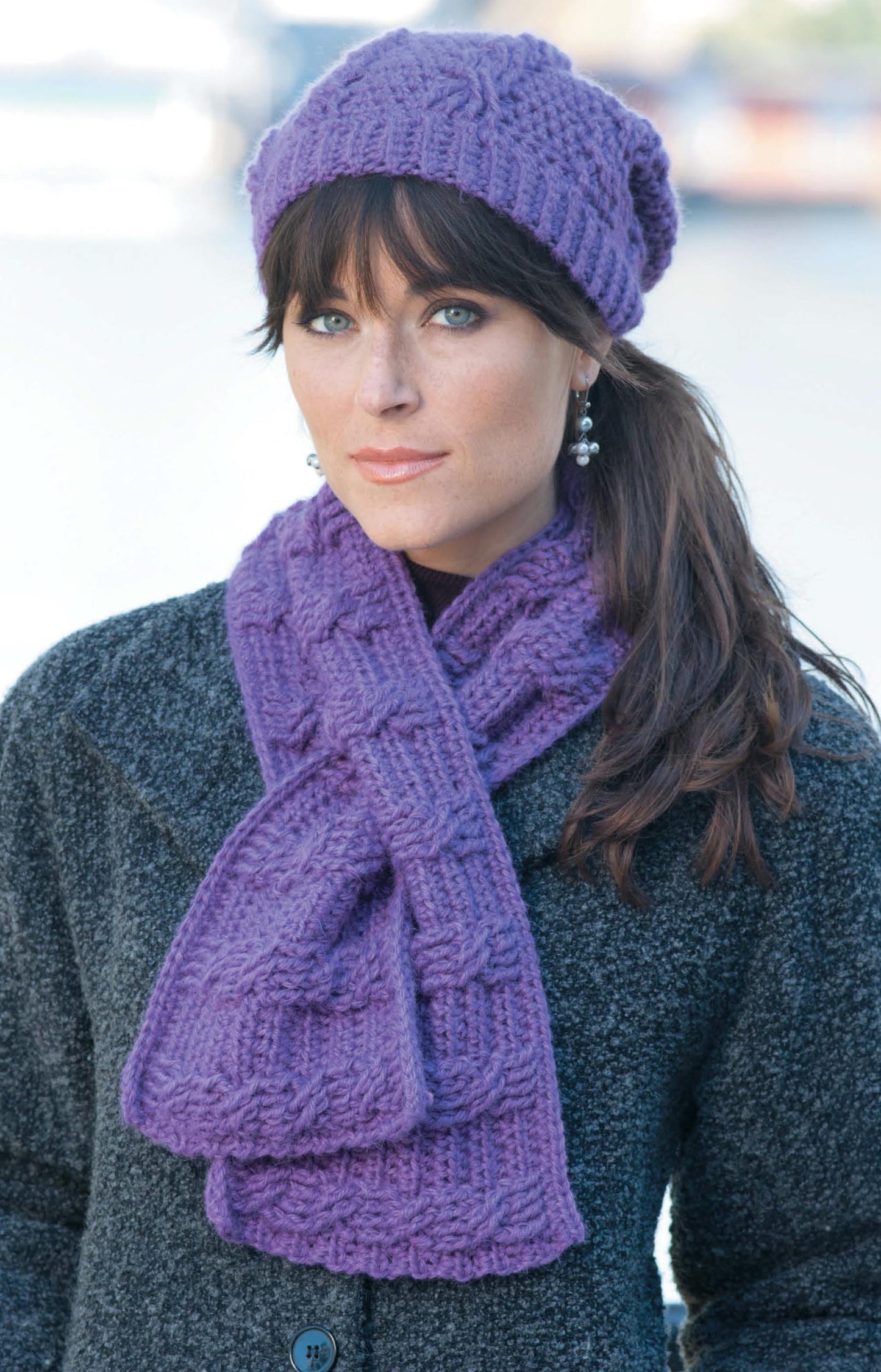 A woman models Leisure Arts' purple Hats & Scarves set over a gray coat, standing outdoors with a neutral expression. The softly blurred background highlights the stylish accessory ensemble.