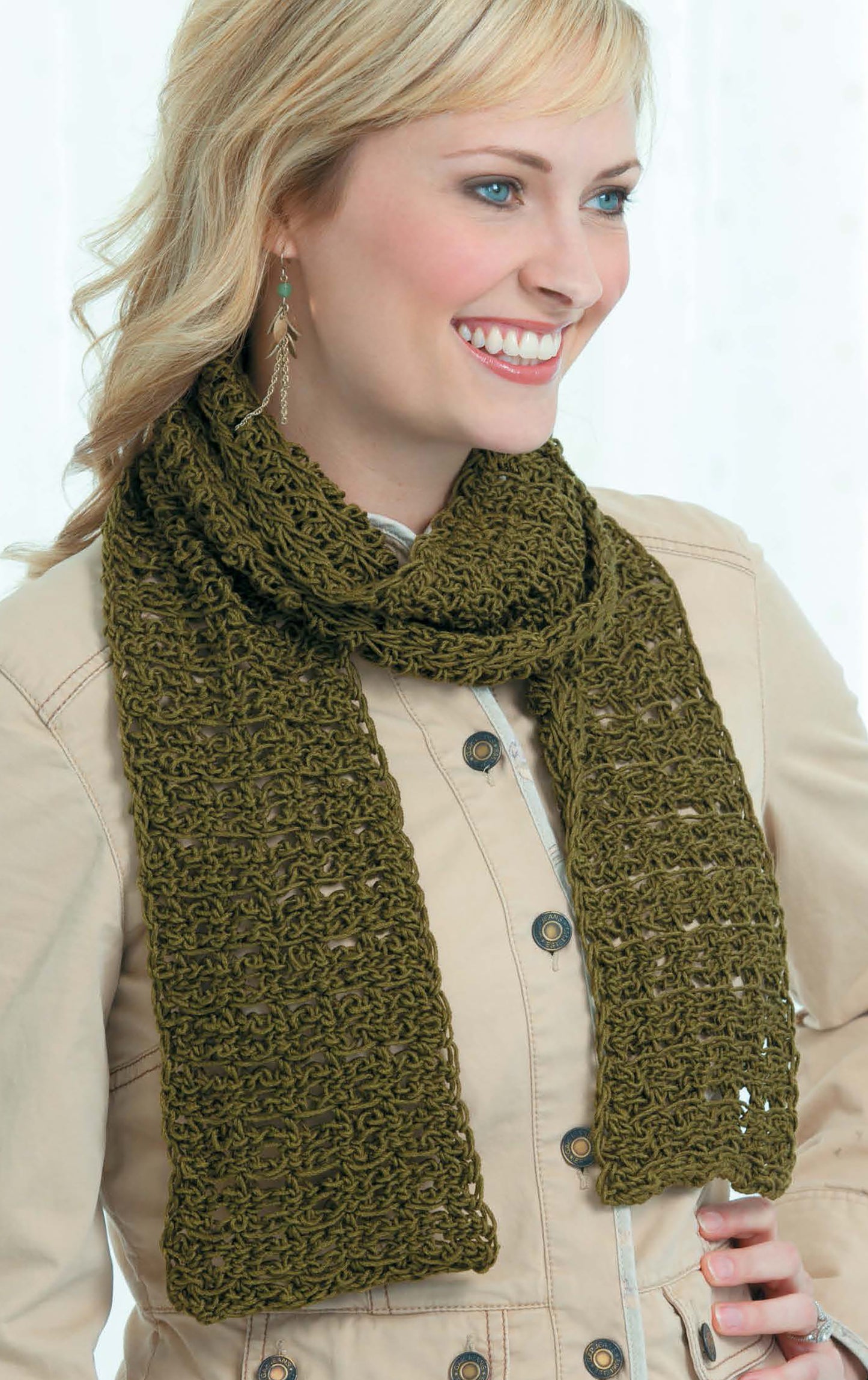 Smiling blonde woman in a tan buttoned jacket and olive green crocheted scarf, highlighting a beginner-friendly design from the "Tunisian Crochet Stitch Guide Digital Download" by Leisure Arts, against a light background.