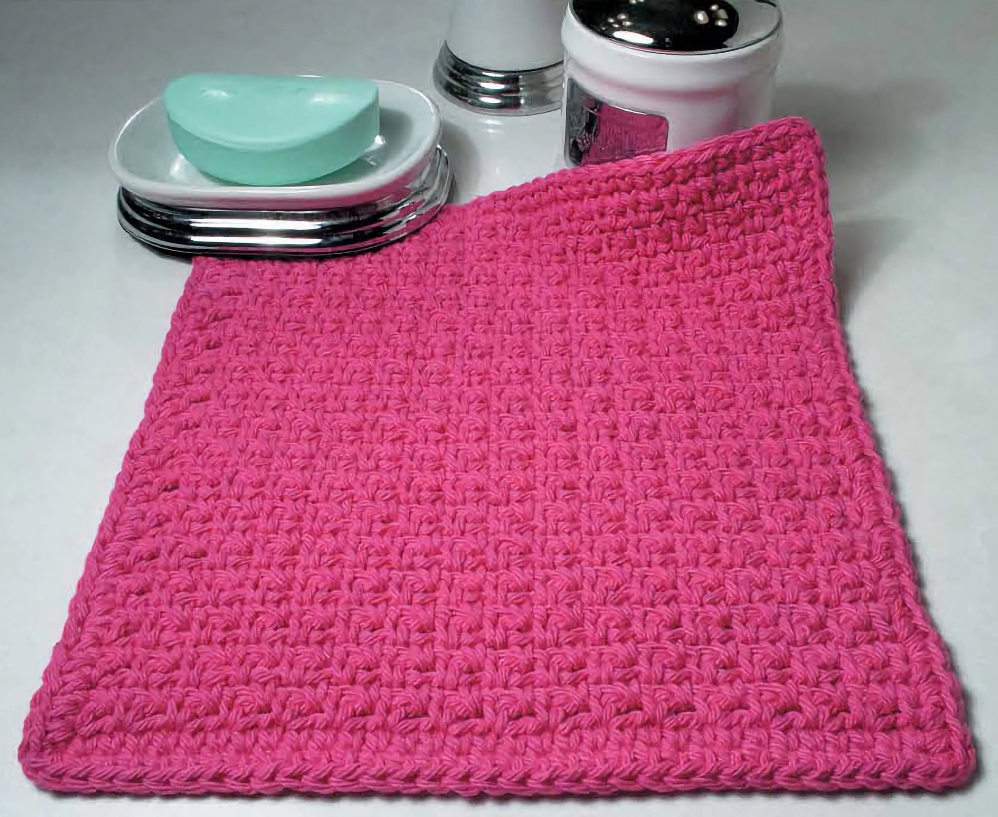 A vibrant pink crocheted washcloth is displayed with a soap dish, green soap, salt shaker, and jar—showcasing Leisure Arts’ "Everything the Internet Didn’t Teach You About Crochet" for stylish and skillful bathroom decor.
