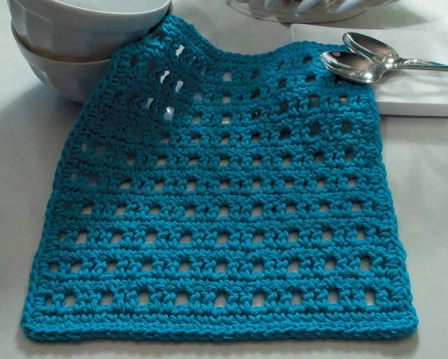 The book "Everything the Internet Didn't Teach You About Crochet" by Leisure Arts sits on a white surface next to stacked white bowls and two metal spoons.