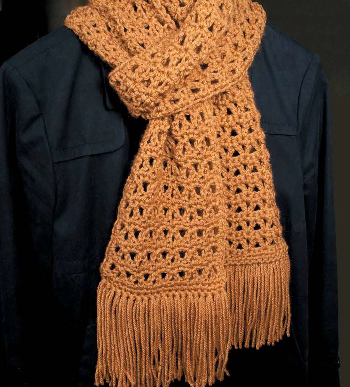 A person in a dark jacket wears a handmade, textured light brown crocheted scarf with fringe—ideal inspiration for anyone using Leisure Arts' "Everything the Internet Didn't Teach You About Crochet" to try new crochet techniques. The scarf is loosely wrapped around the neck.