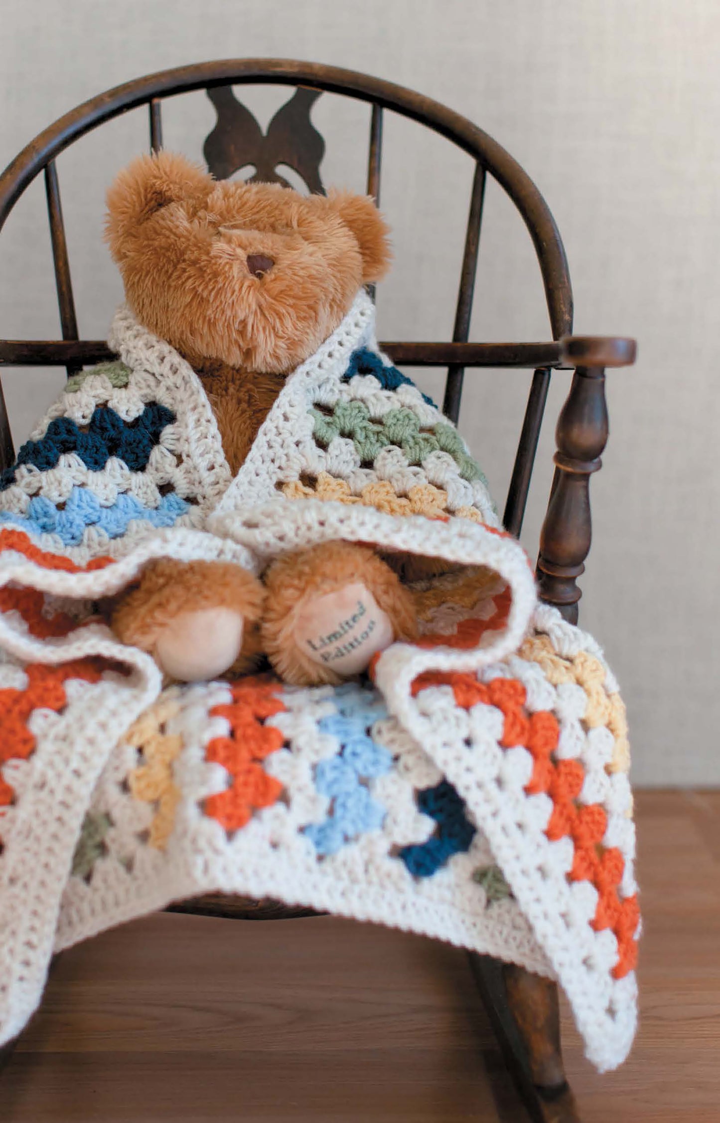 A brown teddy bear sits on a wooden rocking chair, wrapped in a colorful patchwork blanket crocheted from Leisure Arts' Baby Afghans—Cuddly Wraps for Babies & Toddlers! featuring blue, orange, yellow, green, and white squares.