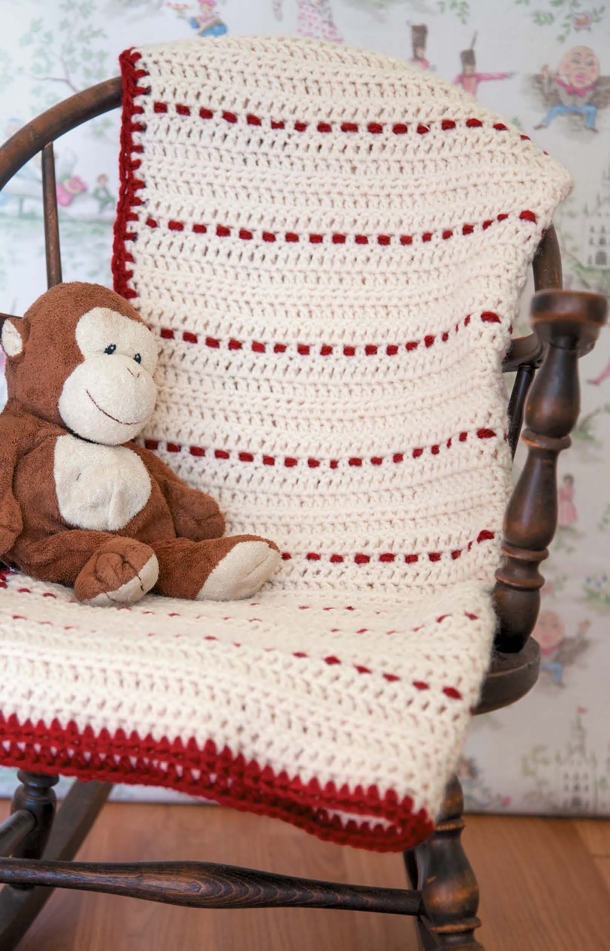 A stuffed brown monkey sits on a rocking chair draped with a Leisure Arts Baby Afghans—Cuddly Wraps for Babies & Toddlers! crocheted blanket in white and red, featuring beginner-friendly patterns. Whimsical wallpaper is in the background.