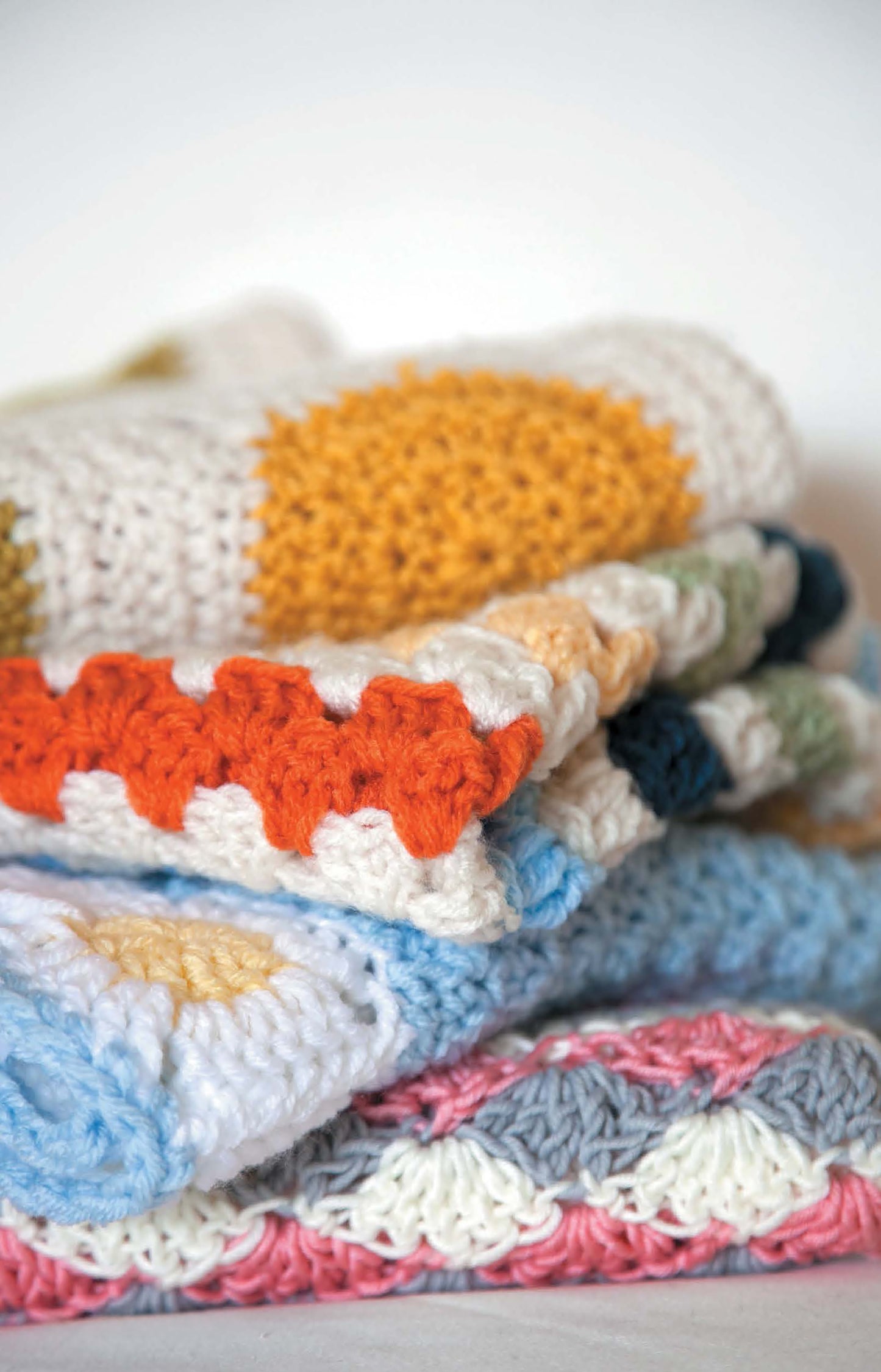 A close-up of folded Baby Afghans—Cuddly Wraps for Babies & Toddlers! by Leisure Arts, showcasing colorful beginner crochet patterns in yellow, orange, blue, pink, gray, and white—perfect for a baby’s nursery.