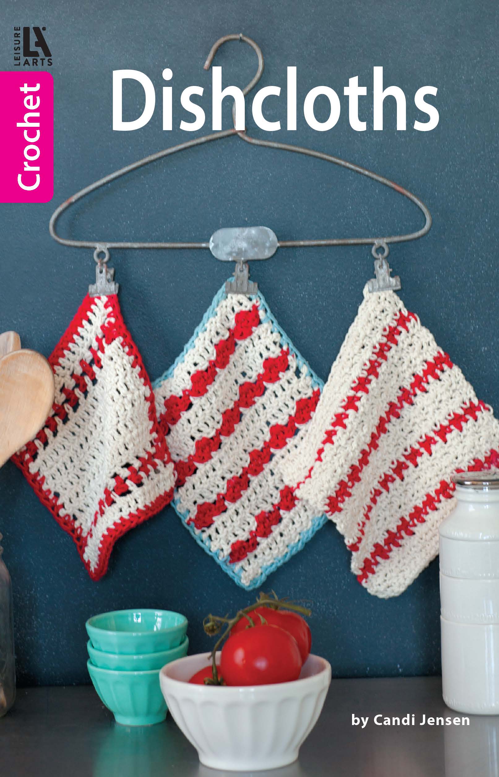 Three crocheted dishcloths in red, white, and cream by Leisure Arts hang from a wire hanger against a dark background. Below are green bowls, a tomato, and a white jar. The text reads Dishcloths by Leisure Arts.