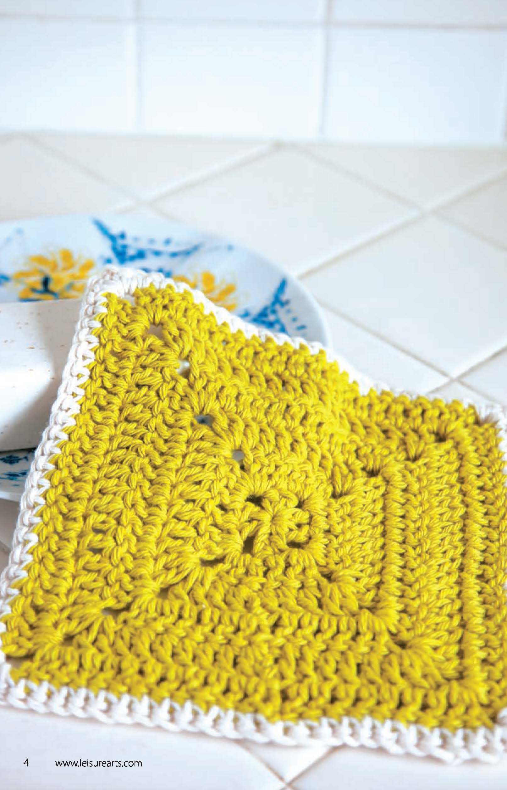 A yellow cotton Leisure Arts Dishcloth with a white border rests on a tiled countertop. A blue and yellow floral plate and a soap bar are partially visible behind it. Ideal for beginner crochet projects.