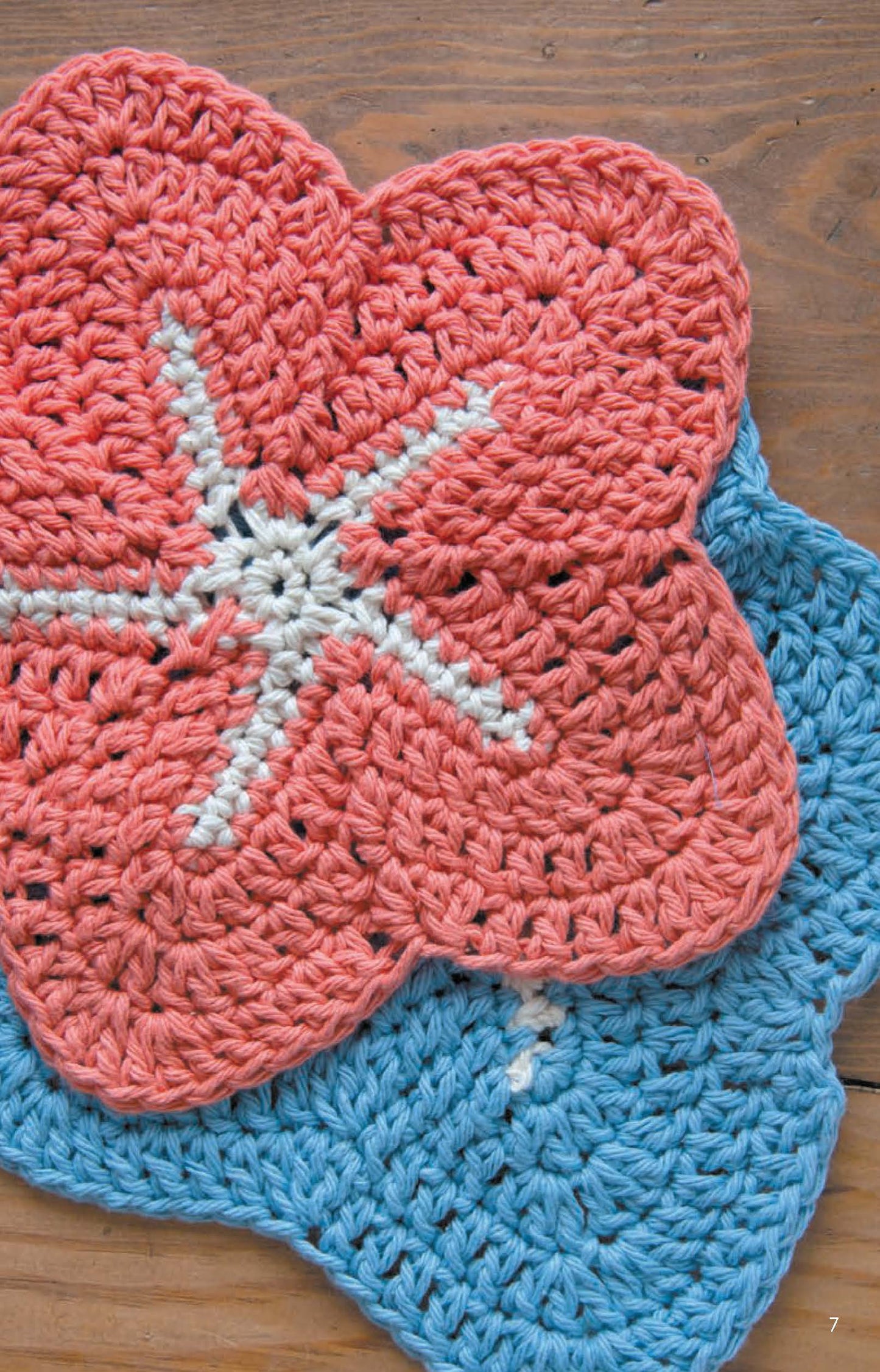 Two Leisure Arts Dishcloths, crocheted from soft cotton yarn, are stacked on a wooden surface. Ideal for beginner crochet projects, one features pink with a white center and the other is blue.