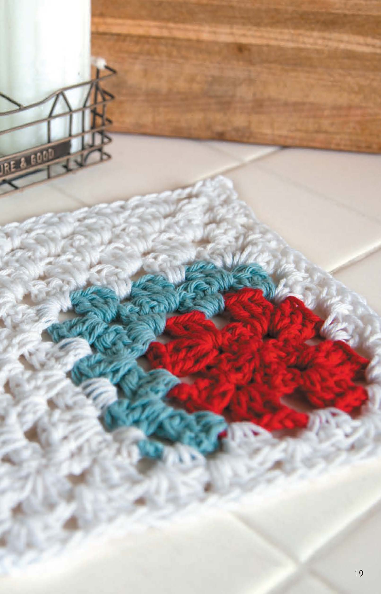 Leisure Arts Dishcloths—crafted in white, teal, and red cotton yarn—sit on a white tiled surface with a candle and wooden box nearby, offering perfect inspiration for beginner crochet projects.