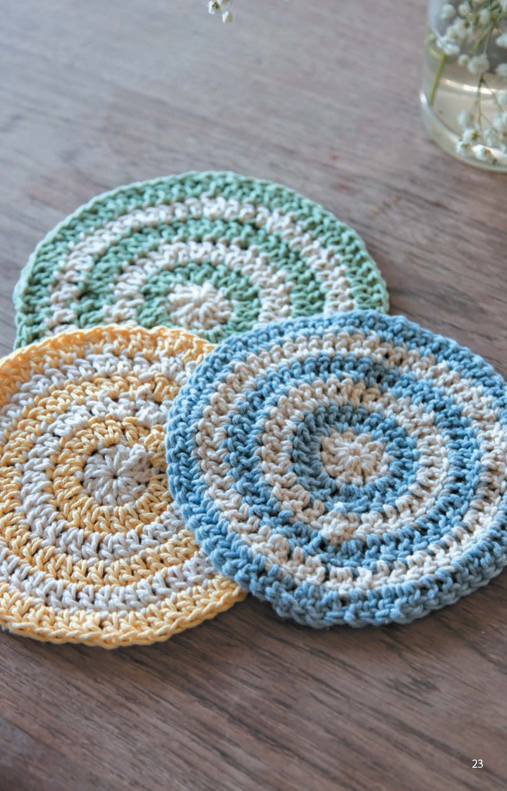 Three Leisure Arts Dishcloths in pastel yellow, green, and blue with white spiral patterns are arranged on a wooden surface. Made from cotton yarn, each dishcloth is round. A vase appears partially in the background.