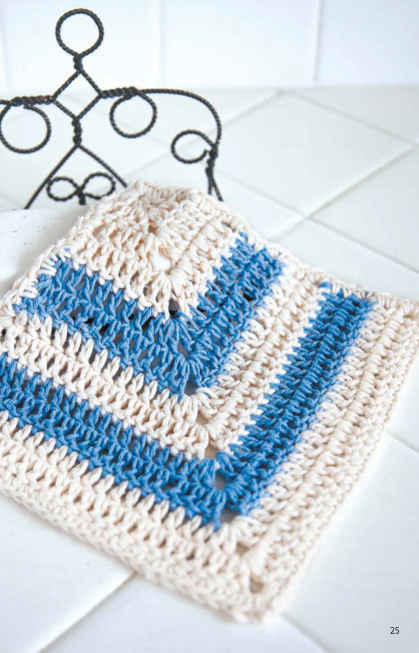 A Leisure Arts Dishcloth in cotton yarn with cream and blue stripes sits on white tiles by a decorative black metal stand—ideal inspiration for beginner crochet projects.
