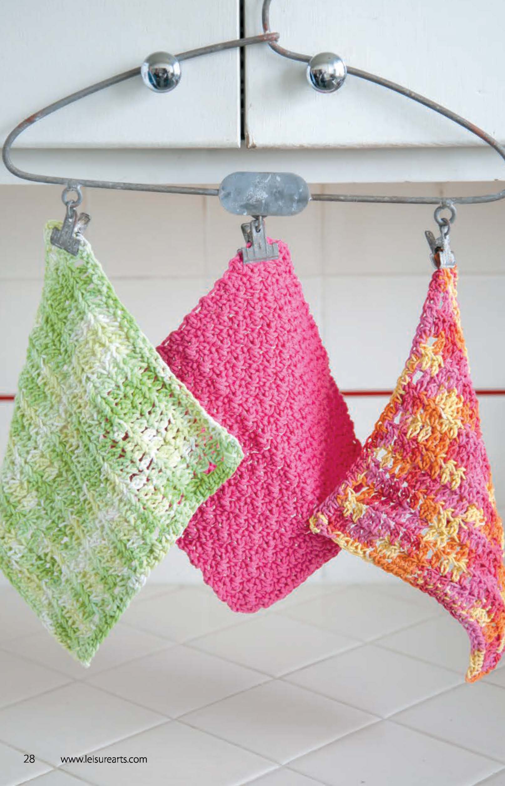 Three Leisure Arts Dishcloths—green and white, solid pink, and pink with orange and yellow—hang from a towel bar on a white cabinet, highlighting beginner crochet projects made from soft cotton yarn.