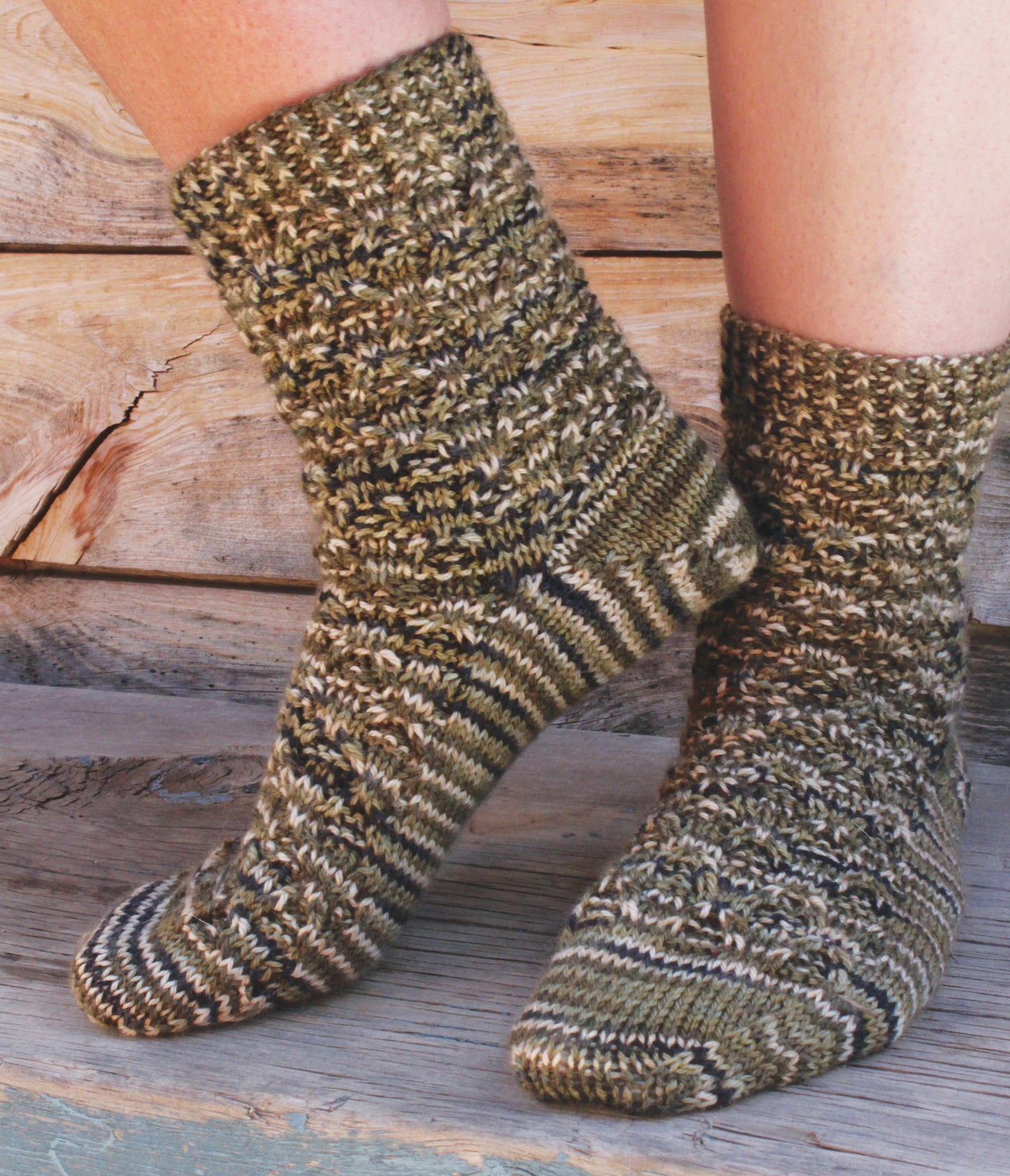 A pair of feet in thick, hand-knit socks featuring a green, brown, and beige variegated pattern—perfect for displaying patterns from Love of Knitting—Socks by Leisure Arts—rest on wooden steps against a rustic wooden backdrop.