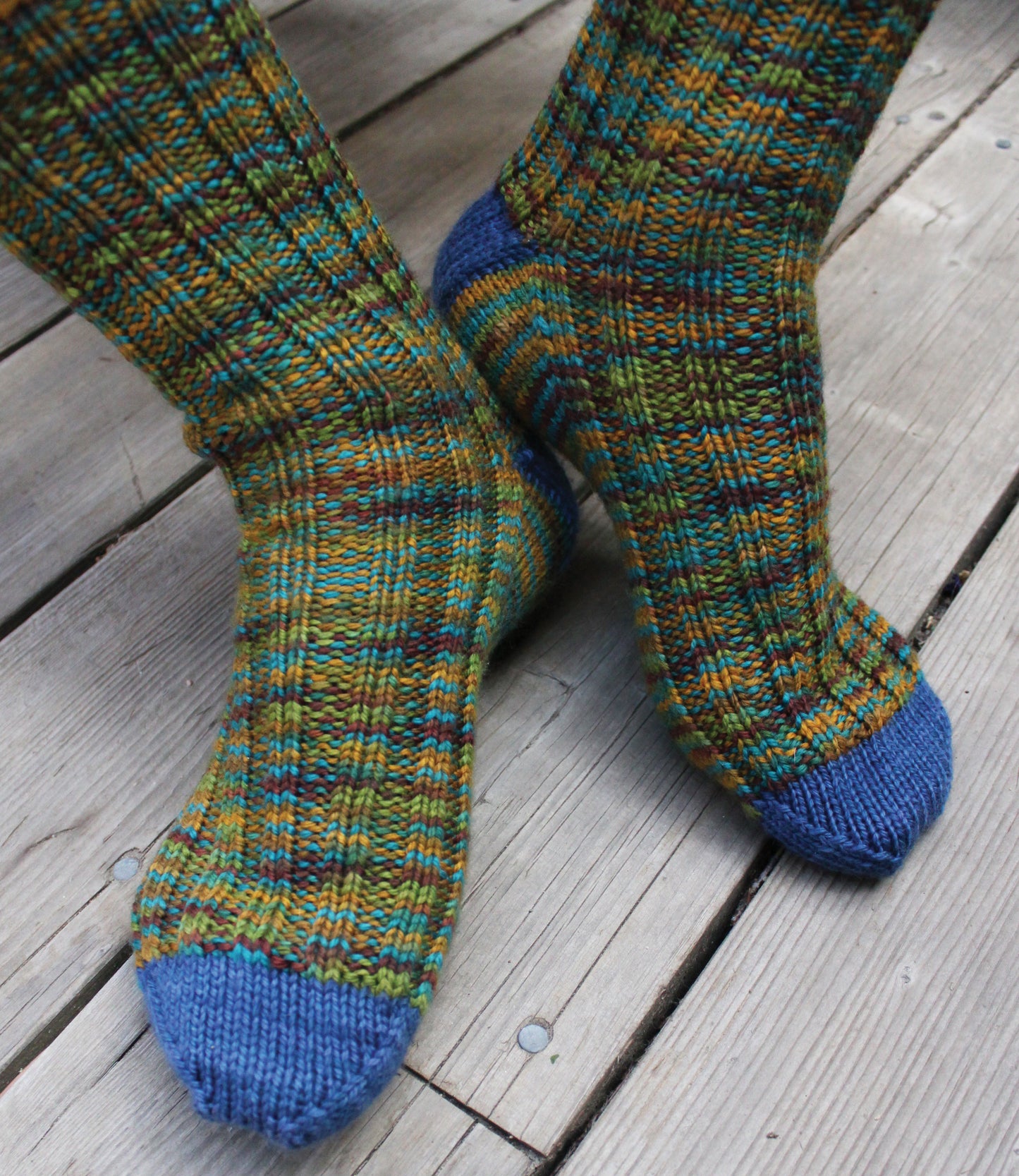 Close-up of feet in cozy hand-knit socks with green, yellow, and blue stripes and solid blue toes and heels on wood floor—perfect inspiration from Leisure Arts’ Love of Knitting—Socks.