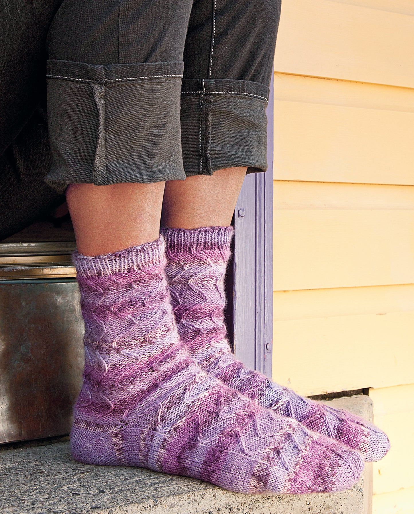 A person wearing purple and white hand-knit socks, inspired by "Love of Knitting—Socks" from Leisure Arts, sits on concrete steps in dark gray cuffed pants near a yellow wooden wall and a purple door frame.