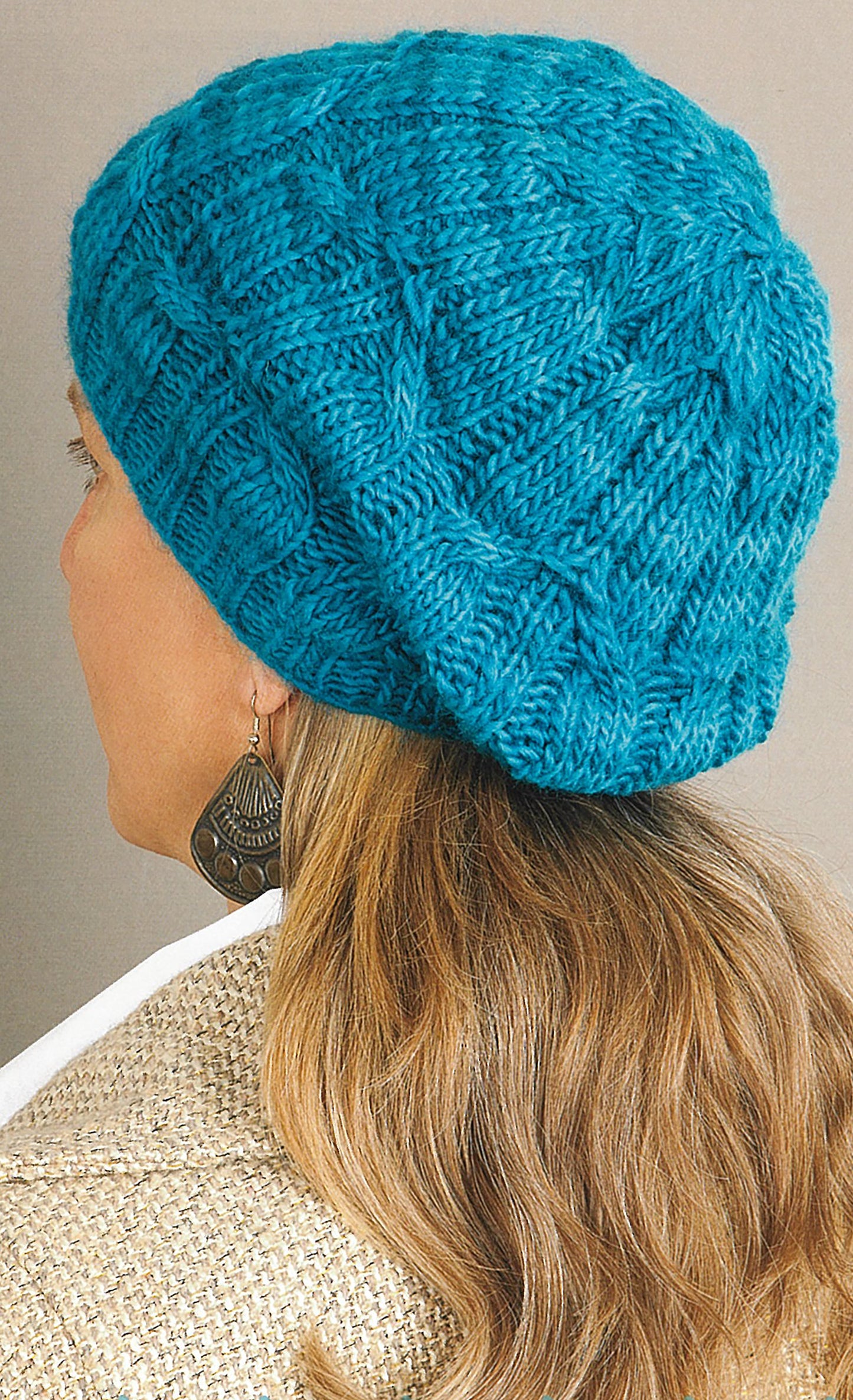 A person with long, wavy blonde hair is seen from behind wearing a bright blue knit hat from Leisure Arts’ "Love of Knitting—Hats & More" collection, a beige textured jacket, and a large patterned dangle earring.