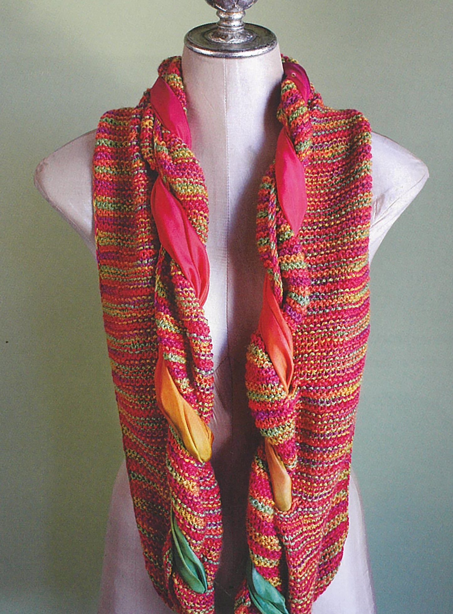 A mannequin displays a vibrant braided scarf in pink, orange, yellow, and green—an ideal beginner project from Leisure Arts' "Love of Knitting—Hats & More" collection—set against a simple light background.