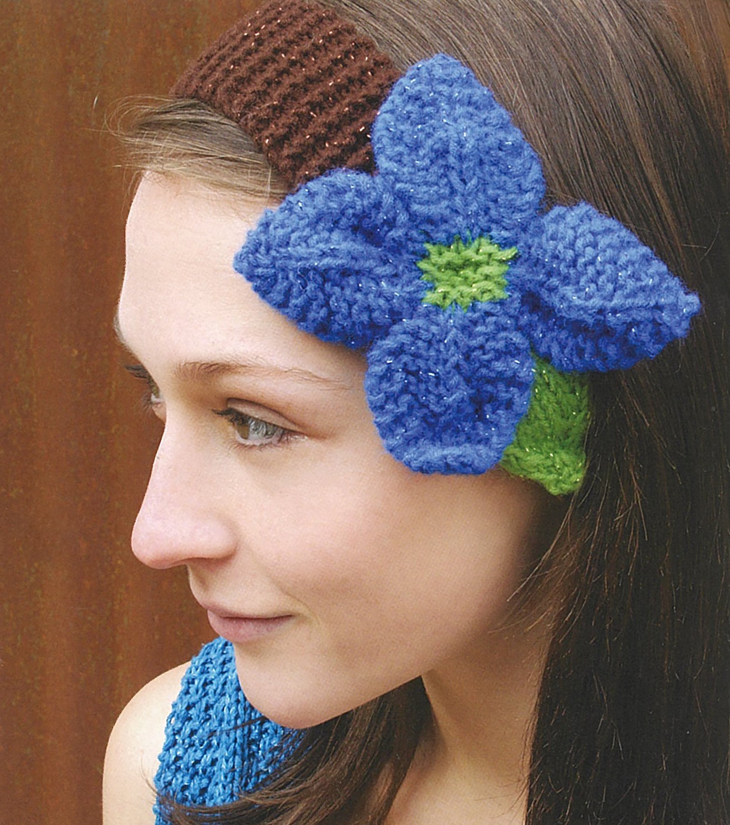 A woman with brown hair wears a beginner-friendly brown knitted headband from Leisure Arts' "Love of Knitting—Hats & More," featuring a large blue flower and green leaves. She looks left, pairing it with a matching blue knit top.