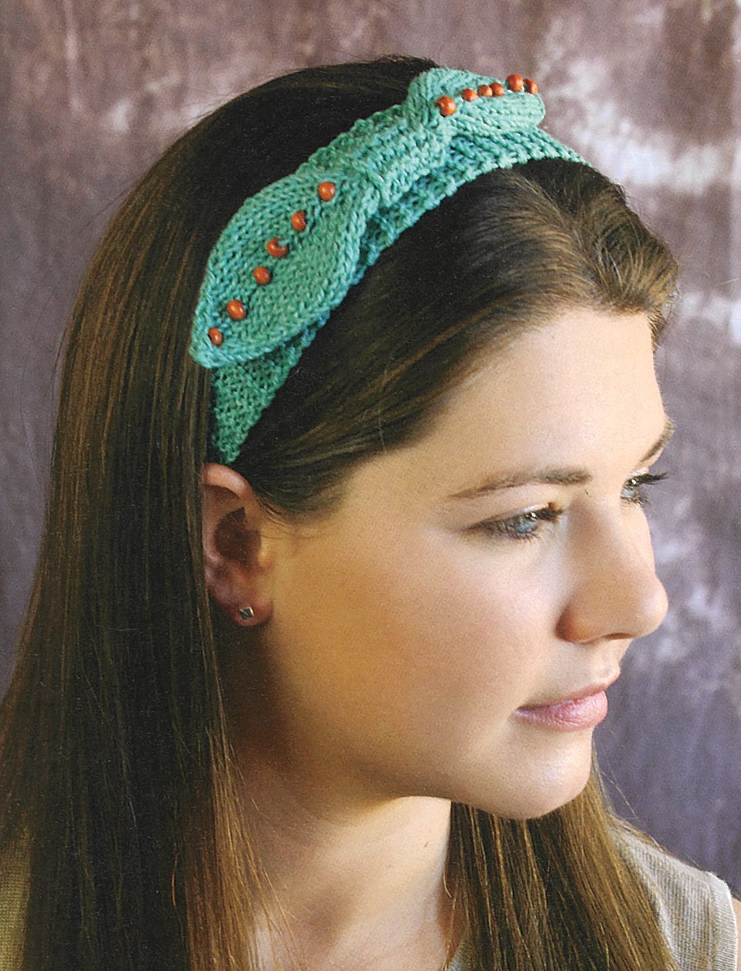 A woman with long brown hair wears a light teal knitted headband from Leisure Arts' Love of Knitting—Hats & More, featuring small orange beads and a front knot, set against a blurred neutral background.