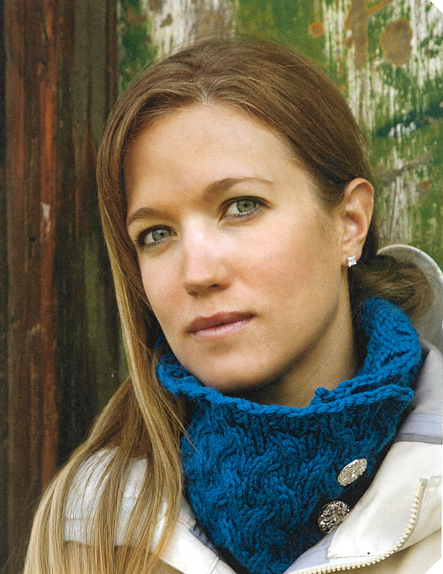A woman with long light brown hair wears a blue knitted neck warmer—an ideal project from Love of Knitting—Hats & More by Leisure Arts—and a light jacket, standing before a rustic wooden backdrop.