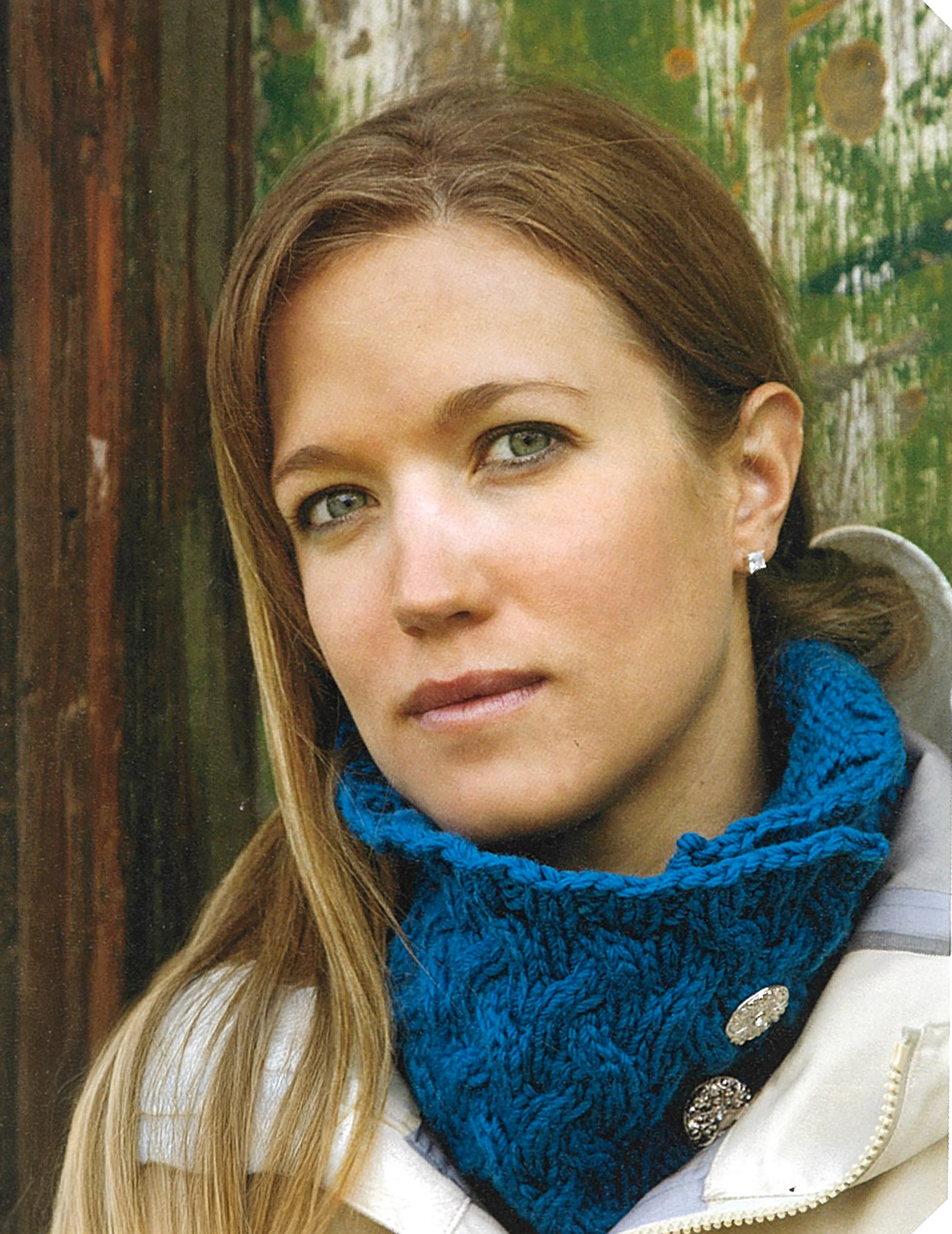 A woman with long light brown hair wears a blue knitted neck warmer—an ideal project from Love of Knitting—Hats & More by Leisure Arts—and a light jacket, standing before a rustic wooden backdrop.