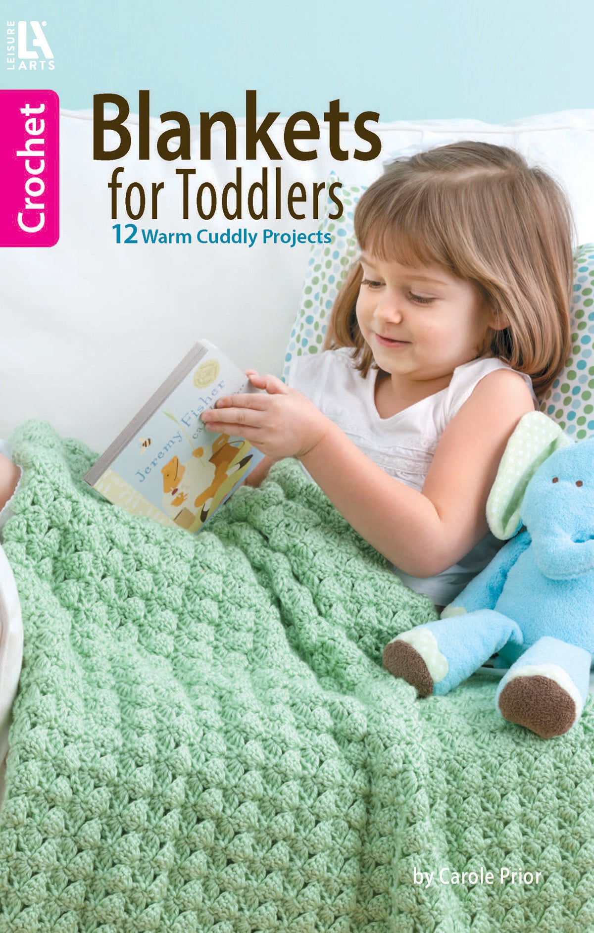 A young girl sits on a bed under the Crochet Blankets for Toddlers by Leisure Arts, holding a book with a blue stuffed animal beside her. Text on the image reads: 12 Warm Cuddly Projects.