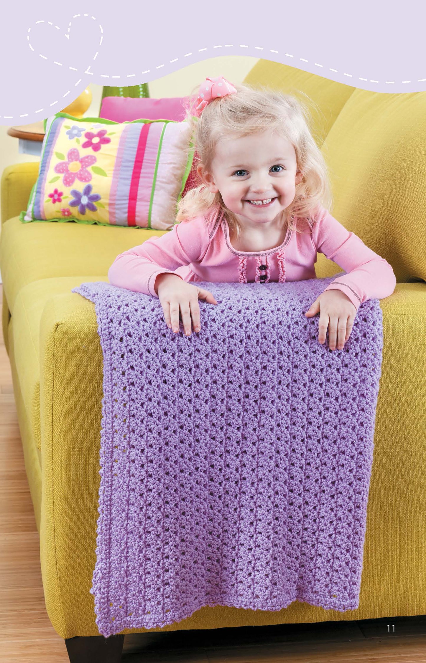 A smiling toddler girl in a pink top leans over a yellow sofa, showing off the Leisure Arts Crochet Blankets for Toddlers in purple. A colorful floral pillow decorates the background.
