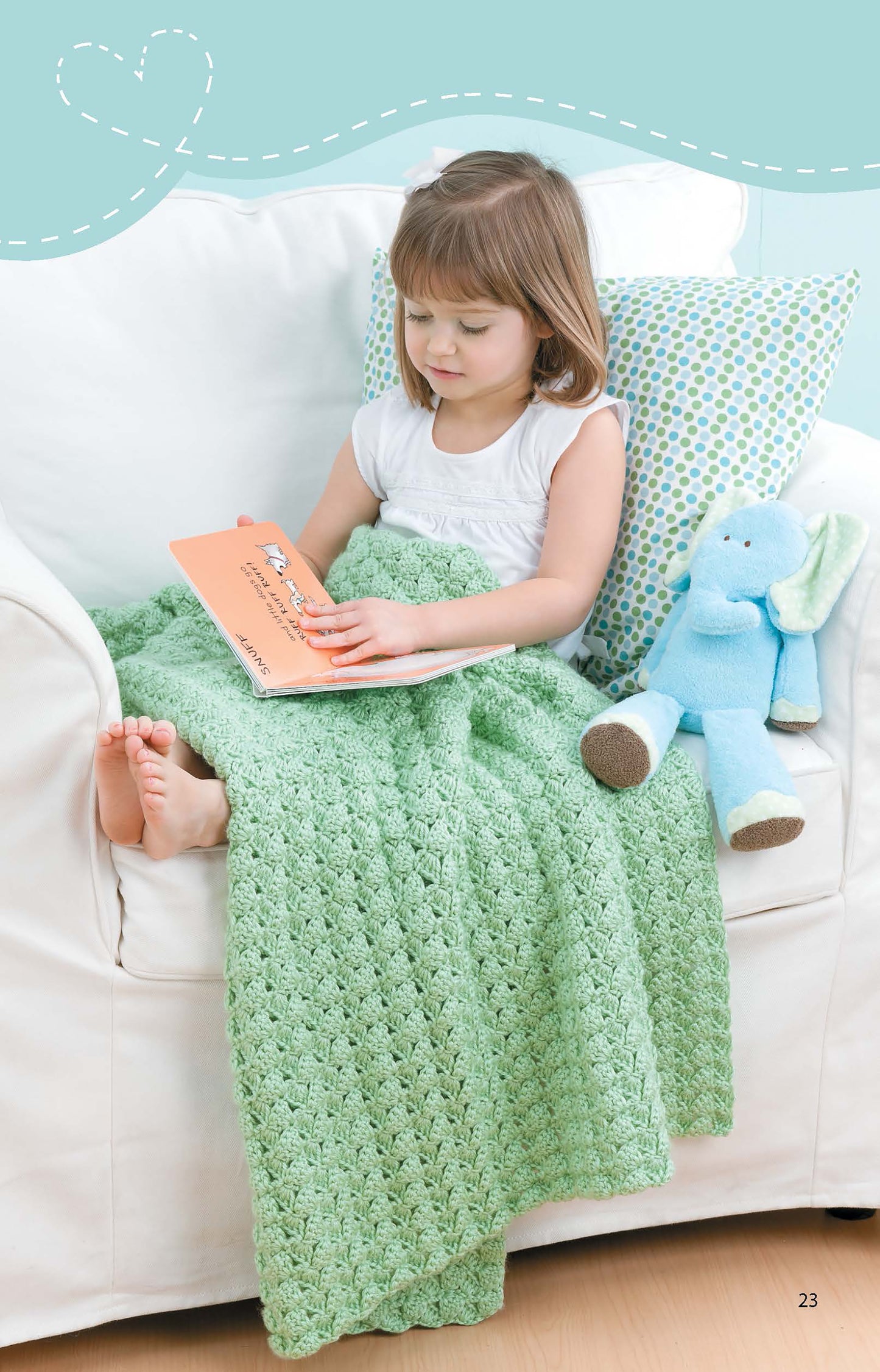 A young girl reads a book on a white couch, covered with a Leisure Arts Crochet Blanket for Toddlers. A blue stuffed elephant toy sits beside her, and there’s a patterned pillow behind her back.