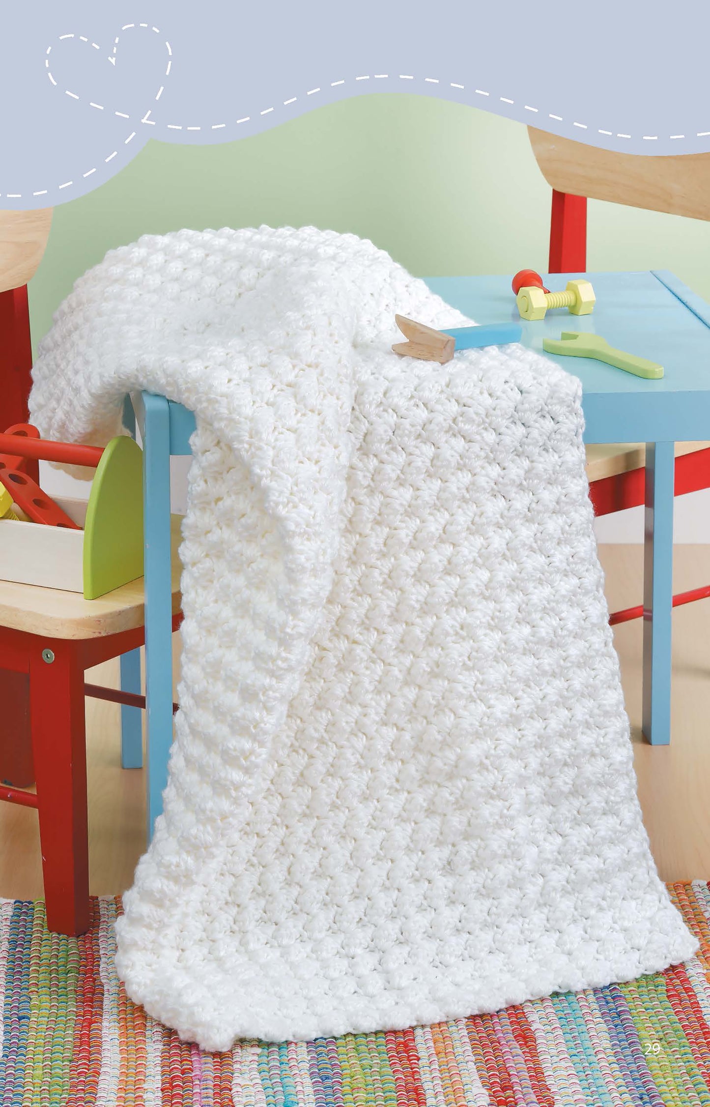 The Leisure Arts Crochet Blankets for Toddlers, in a textured white knit made from medium weight yarn, is draped over a small blue table between two wooden chairs with red legs; a colorful toy and rug are also visible.