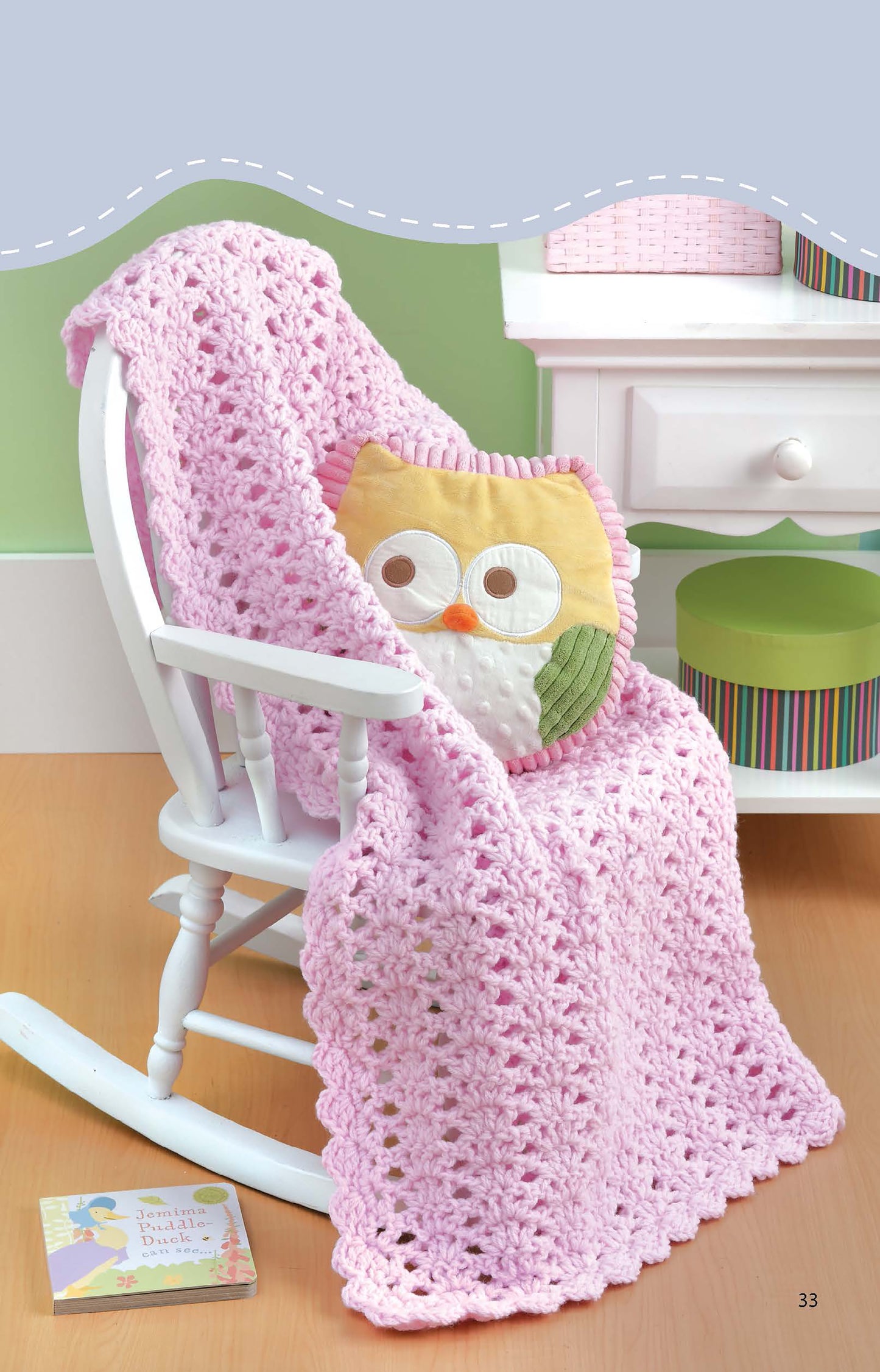 A white rocking chair draped with a Leisure Arts Crochet Blankets for Toddlers, paired with an owl pillow, sits by a white table holding a green box and pink basket. An open children’s book is on the floor nearby.