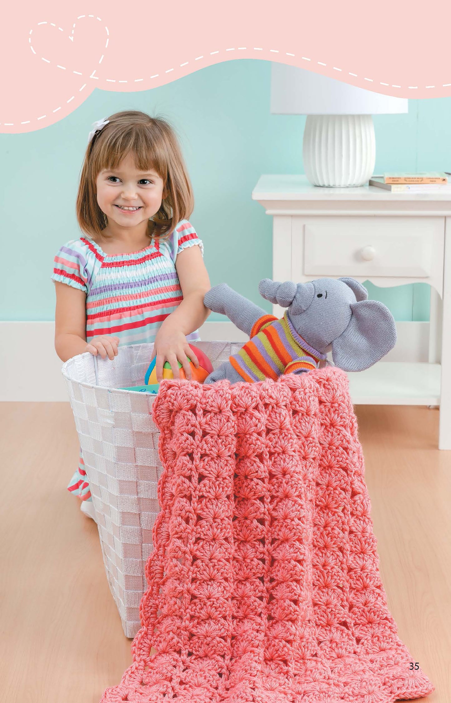 A young girl smiles while placing a plush elephant and colorful balls into a basket topped with a Leisure Arts Crochet Blanket for Toddlers made from medium weight yarn. A white nightstand and lamp sit in the background.