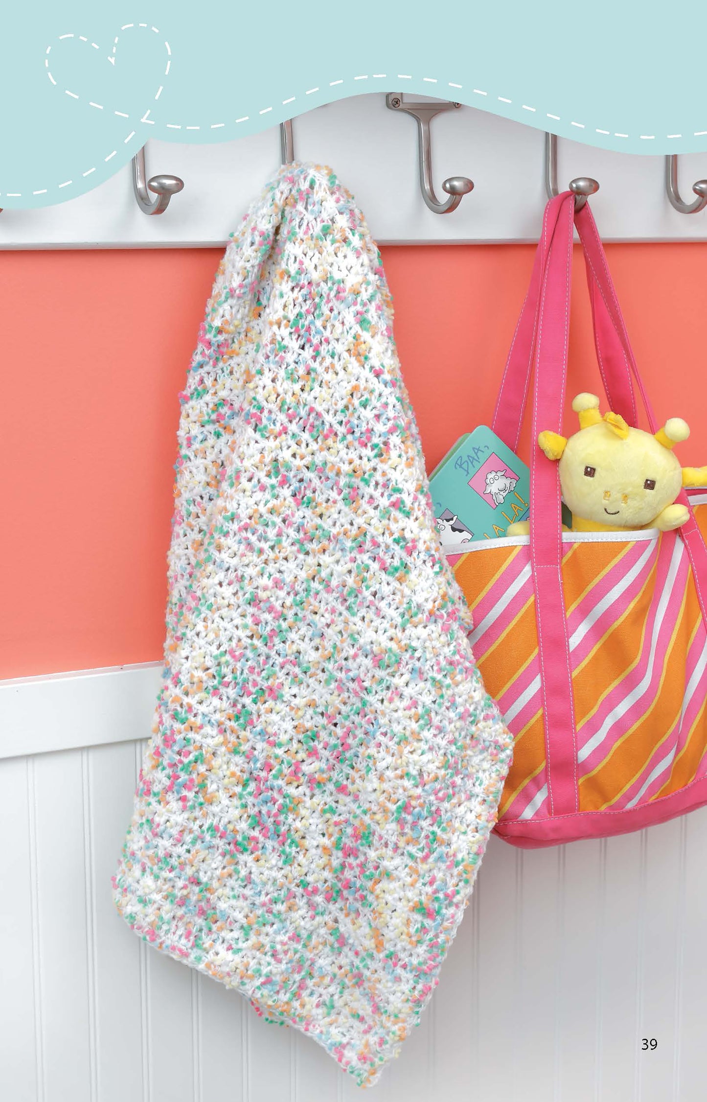 A pastel multicolored "Crochet Blankets for Toddlers" by Leisure Arts hangs on a hook near a pink-orange striped bag with a yellow plush toy and blue children’s book, set against a coral wall with white wainscoting.