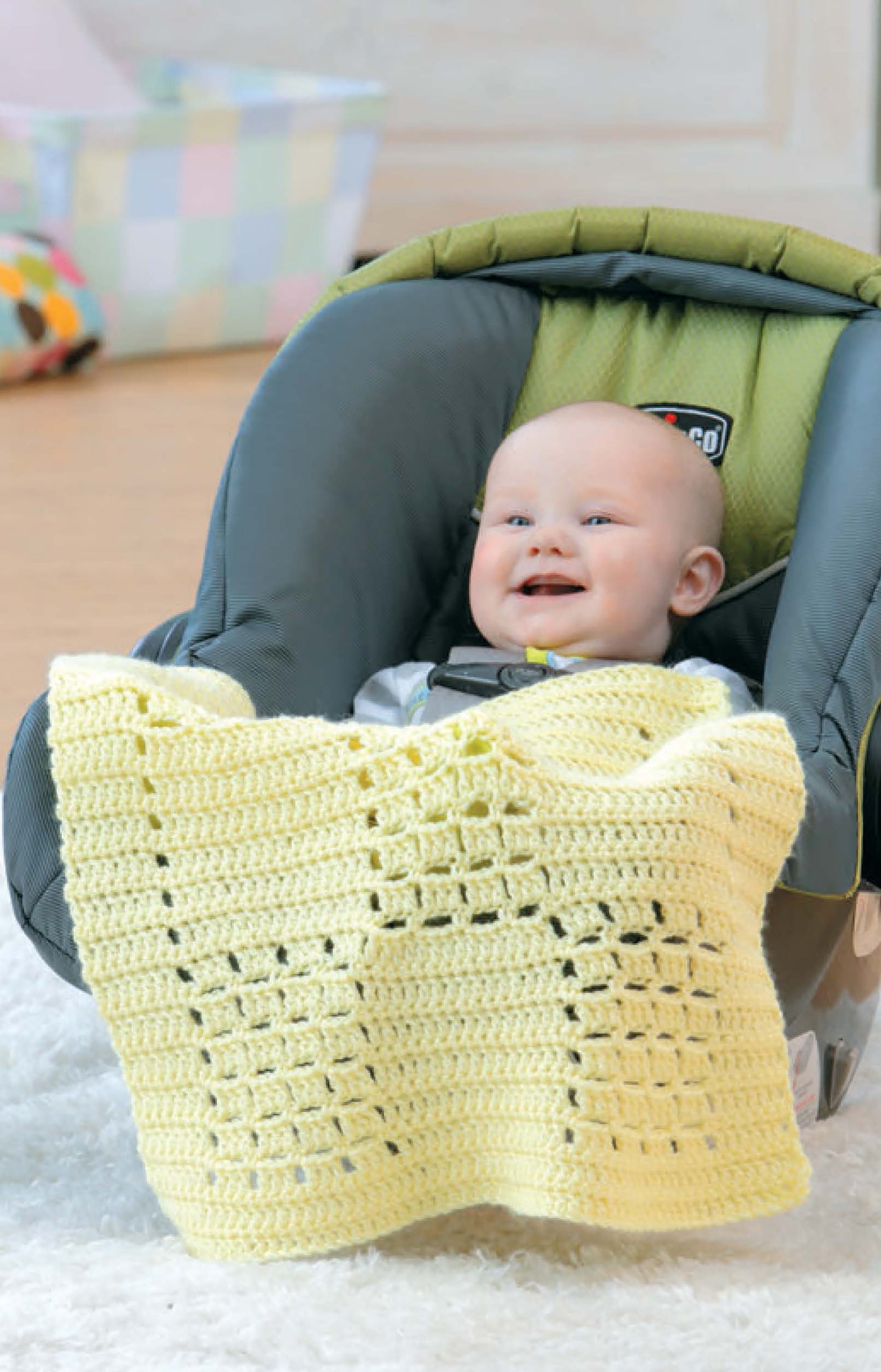 A smiling baby sits in a car seat with a light yellow Leisure Arts Crochet Car Seat Blanket, a perfect beginner-friendly pattern and thoughtful baby shower gift, displayed on a soft white rug indoors.