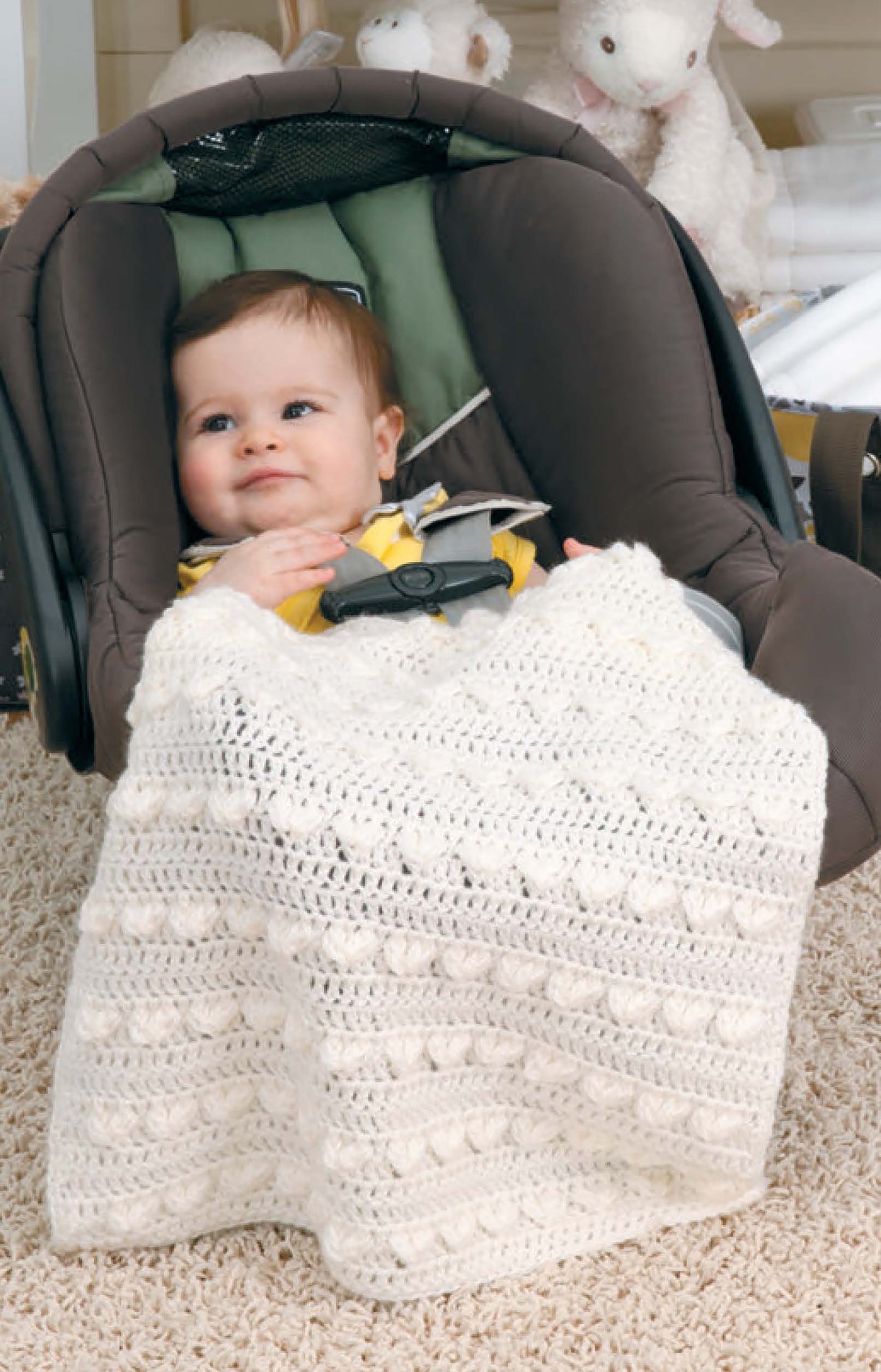 A smiling baby sits in a car seat with a Leisure Arts Crochet Car Seat Blanket—an ideal baby shower gift. Plush stuffed animals and nursery items appear softly blurred in the background.