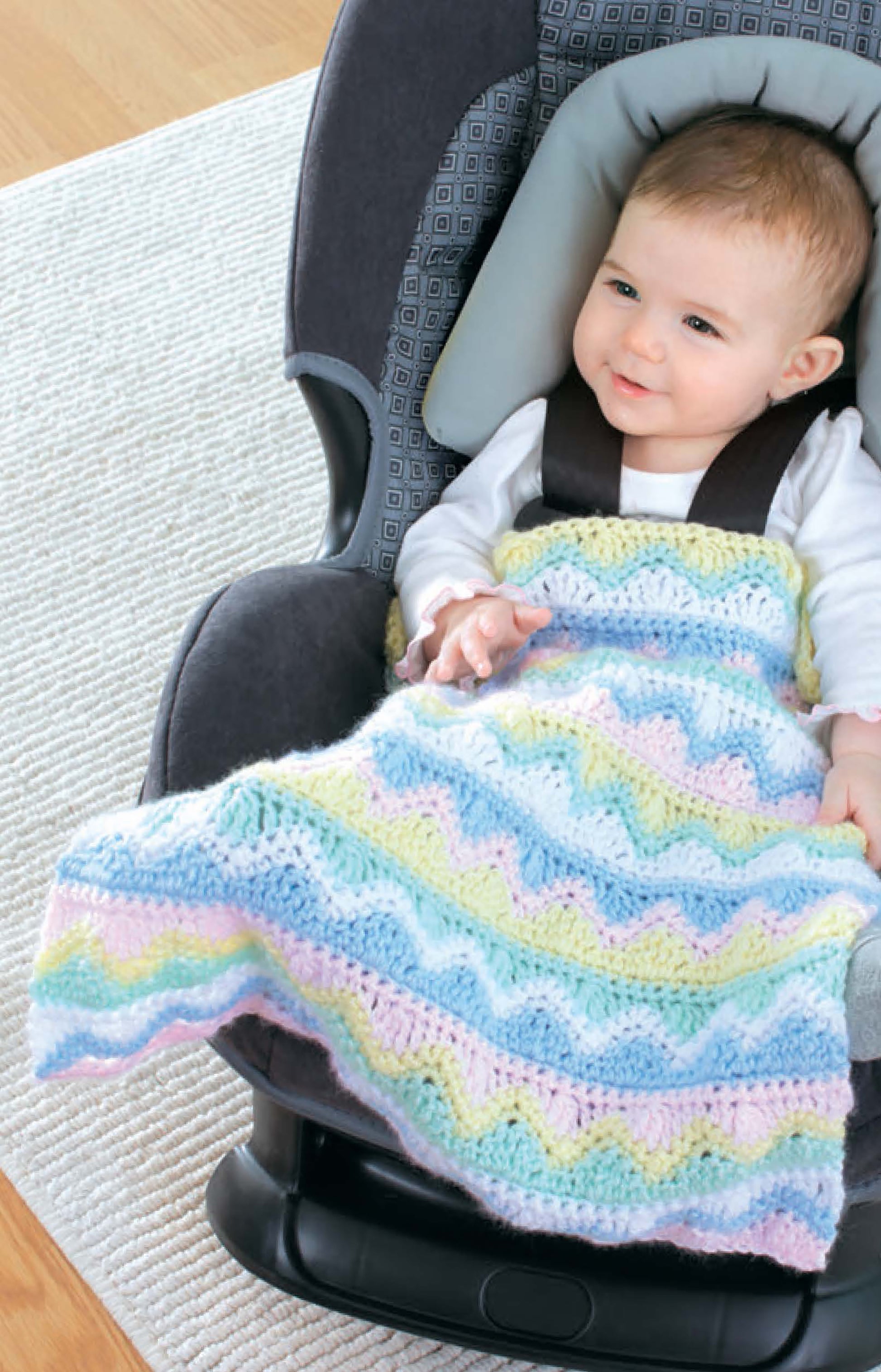 A smiling baby sits in a car seat, wrapped in a Crochet Car Seat Blanket by Leisure Arts featuring colorful pastel wavy stripes—an easy crochet blanket and perfect baby shower gift. A light-colored rug lies on the floor beside the seat.