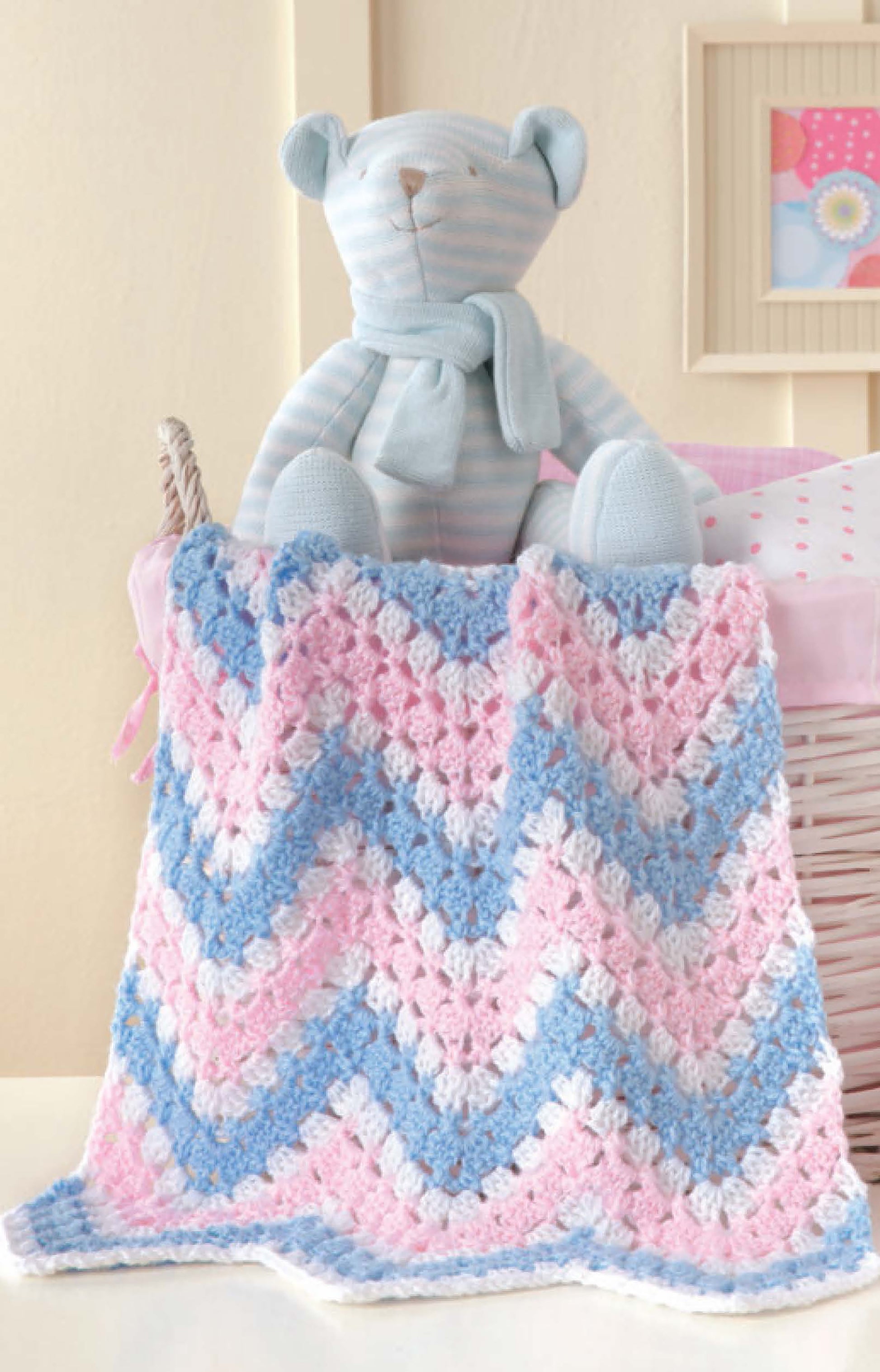 A blue and white striped teddy bear sits in a basket with a Leisure Arts Crochet Car Seat Blanket in pink, blue, and white draped over the edge—perfect for baby shower gifts. A framed picture and part of a pink pillow are in the background.