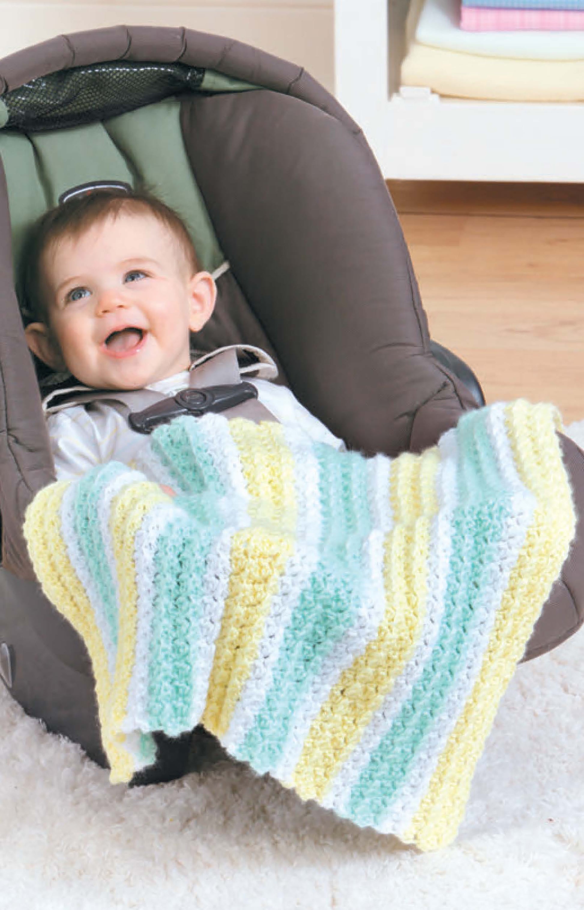 A smiling baby sits in a car seat, wrapped in a Leisure Arts Crochet Car Seat Blanket with pastel stripes—ideal for baby shower gifts. Neatly folded towels and books fill the shelves in the background.