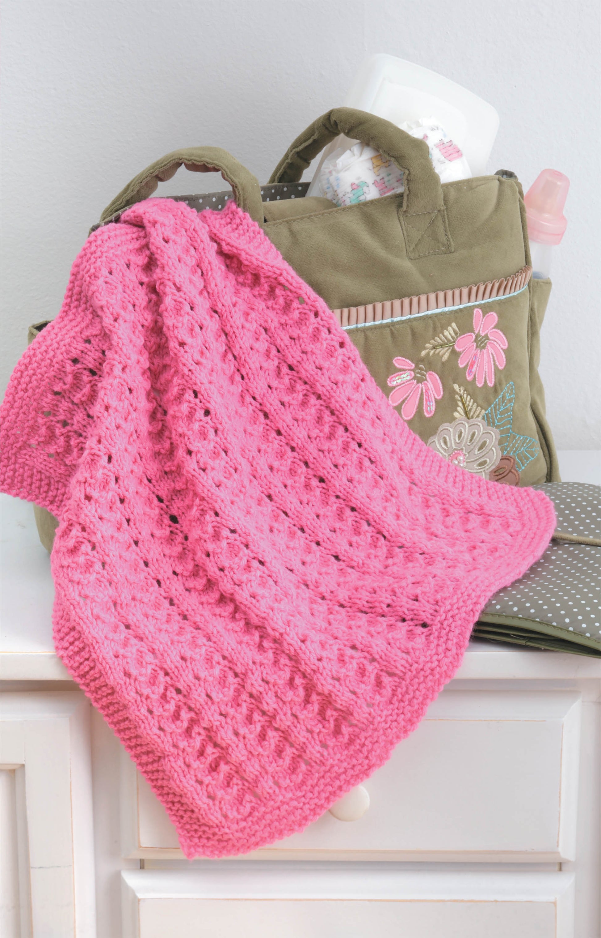 A green diaper bag with floral embroidery sits on a white dresser. Draped over it is a pink Leisure Arts “Knit Car Seat Blankets” digital download—perfect for easy knitting projects or baby shower gifts—with baby items like a bottle, wipes, and diapers nearby.