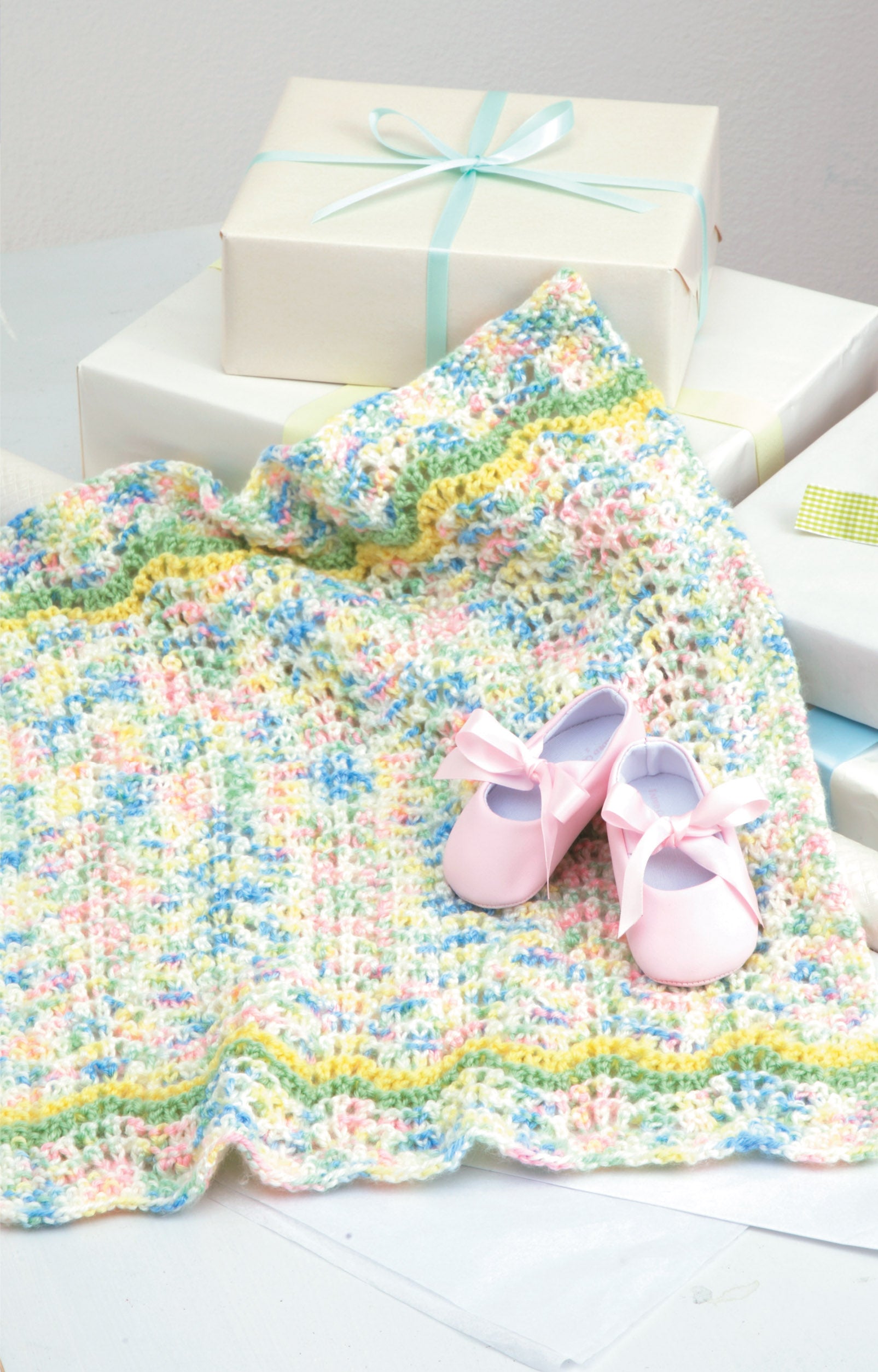 The Leisure Arts Knit Car Seat Blankets Digital Download is a charming and easy knitting project, perfect for creating thoughtful baby shower gifts. Display it with pink baby shoes near white gift boxes tied with blue ribbon.