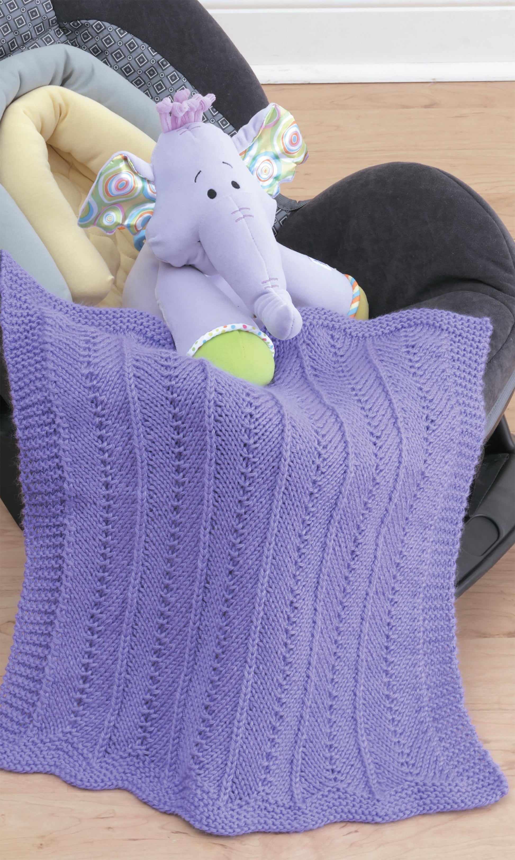 The Leisure Arts Knit Car Seat Blankets Digital Download makes a charming baby shower gift, especially when draped over a car seat with a plush elephant toy featuring colorful patterned ears and a purple crown.