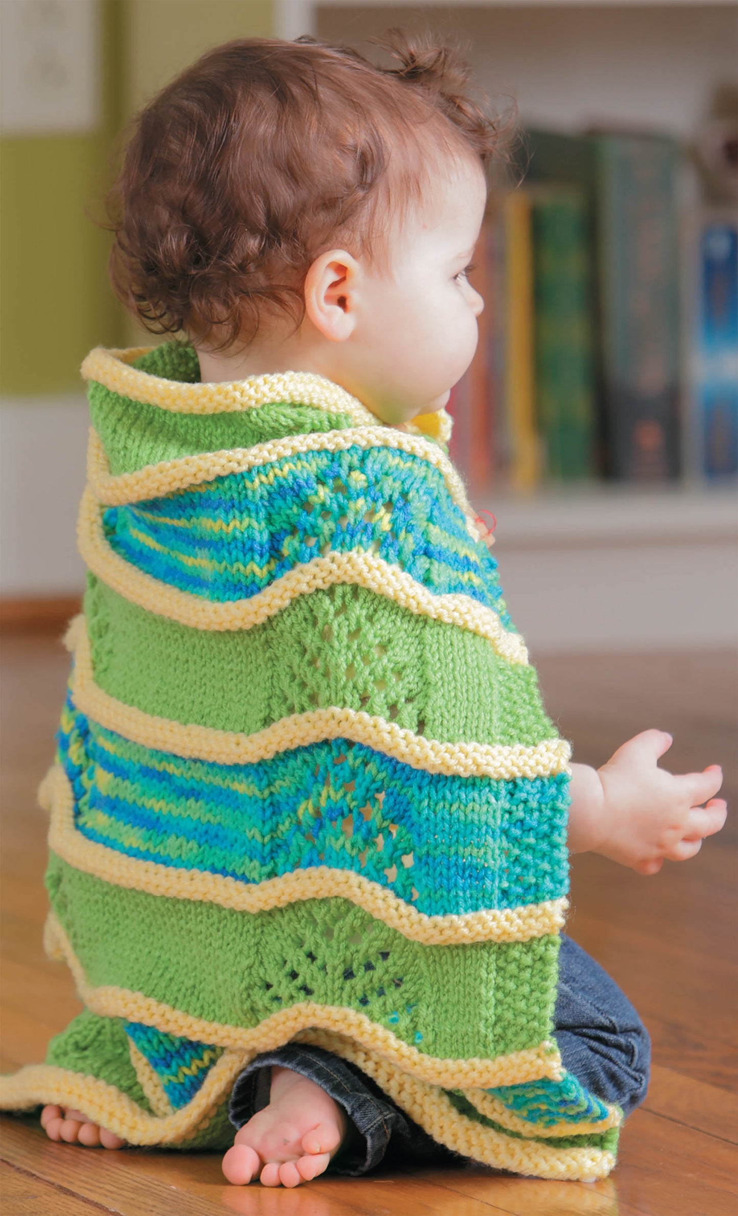 A baby with curly hair sits on the floor wrapped in a green, yellow, and blue striped blanket from Leisure Arts’ Knit Car Seat Blankets Digital Download—quick to knit and perfect as a baby shower gift. Books are visible on a shelf in the background.