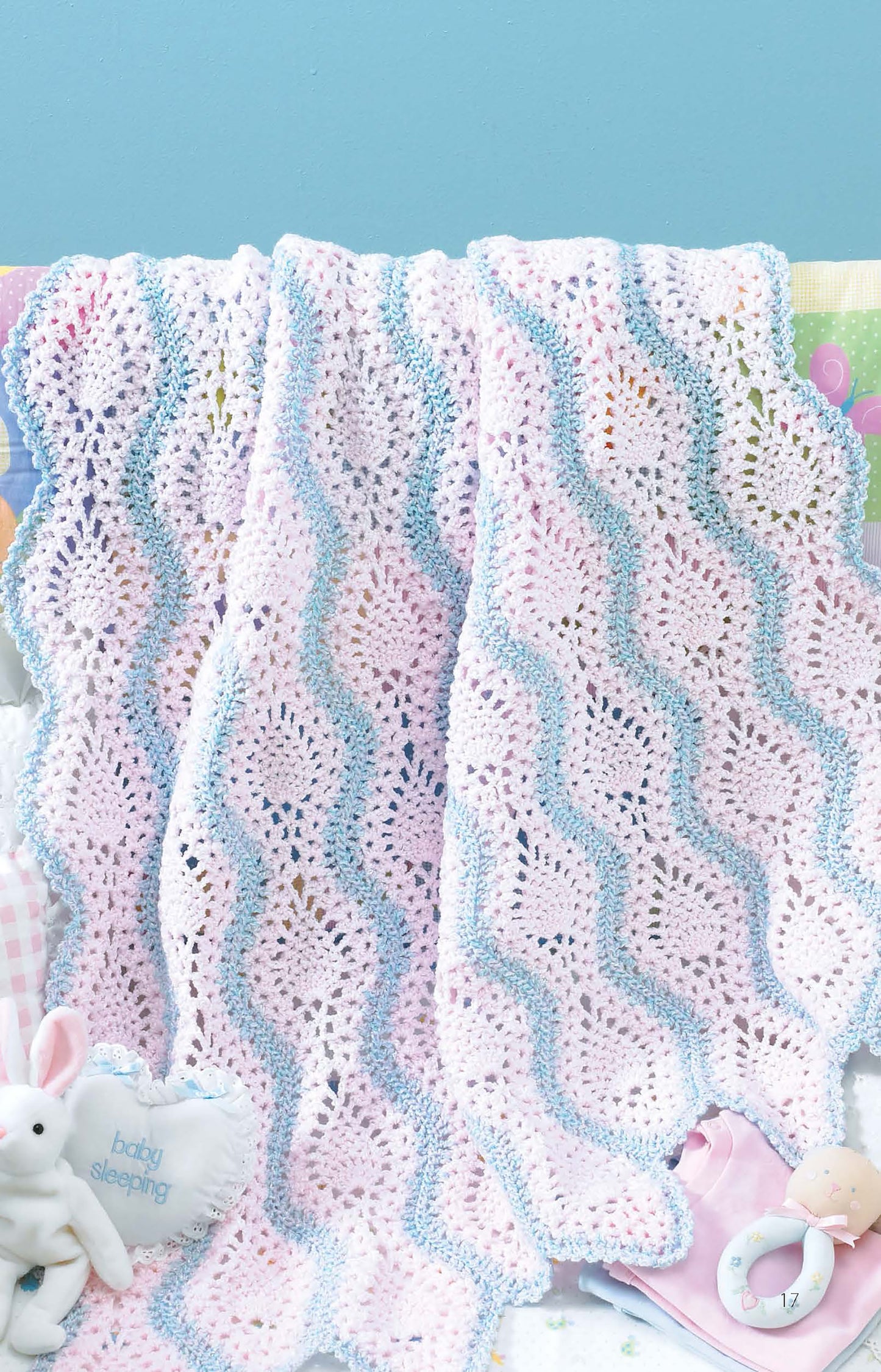 A pastel baby blanket made using the “Learn to Make Mile-a-Minute Baby Afghans” digital download by Leisure Arts, with wavy pink, white, and blue stripes, is draped over a crib rail near soft toys and a “baby sleeping” pillow.