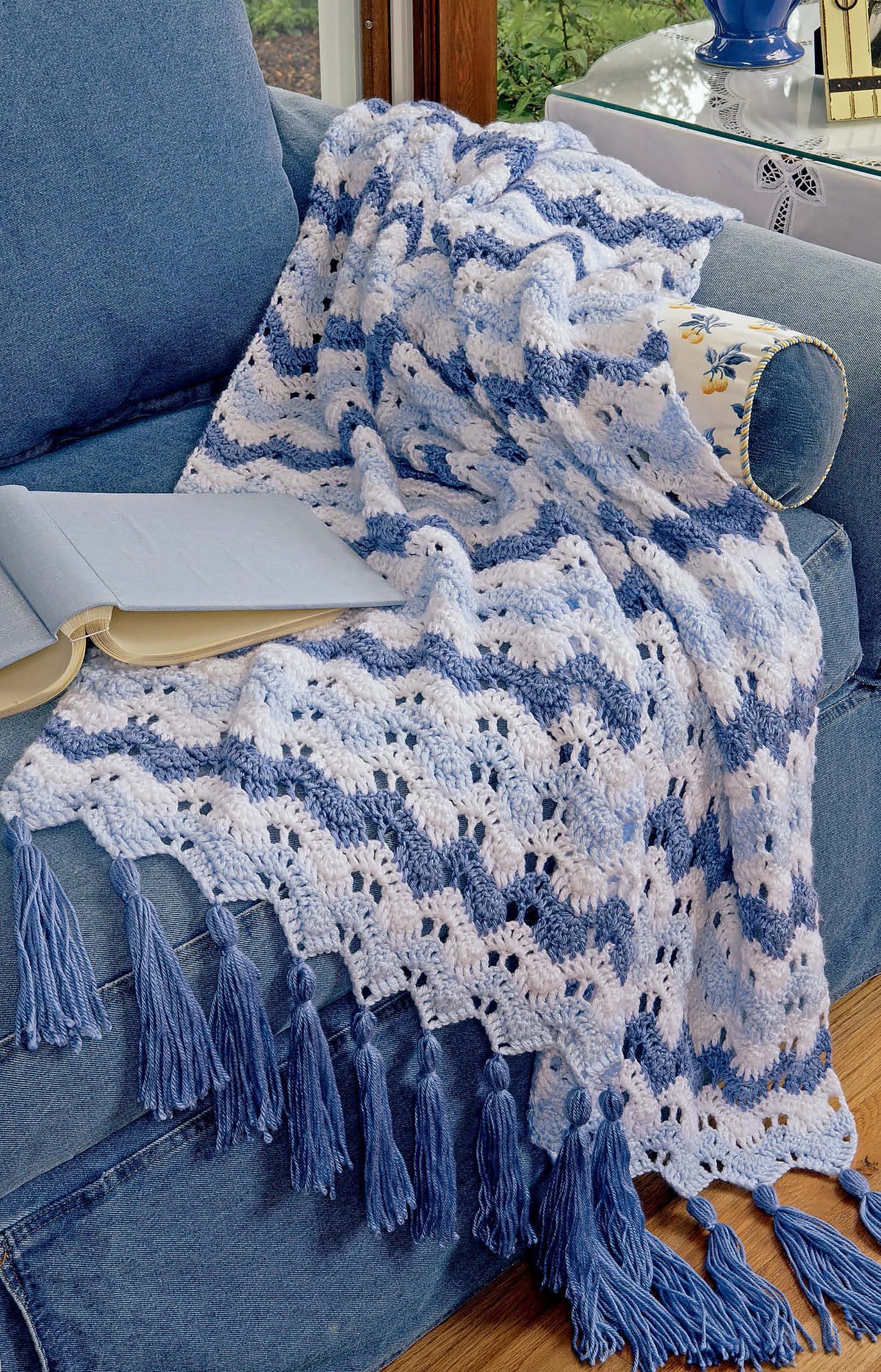 A blue and white striped blanket inspired by Leisure Arts’ Learn to Crochet Ripple Afghans Digital Download drapes over a blue sofa. Sunlight streams in as an open book sits on the arm of the sofa.