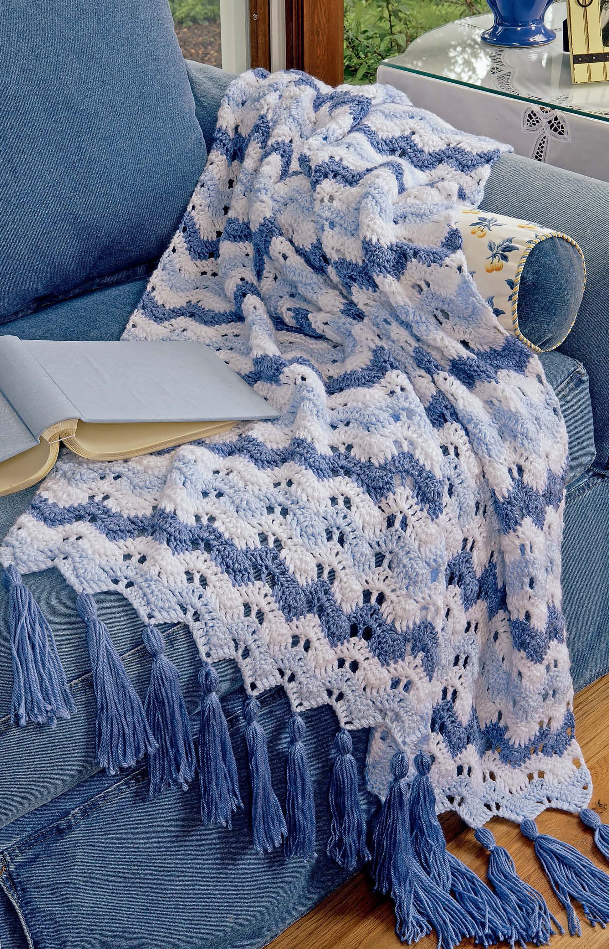 A blue and white striped blanket inspired by Leisure Arts’ Learn to Crochet Ripple Afghans Digital Download drapes over a blue sofa. Sunlight streams in as an open book sits on the arm of the sofa.