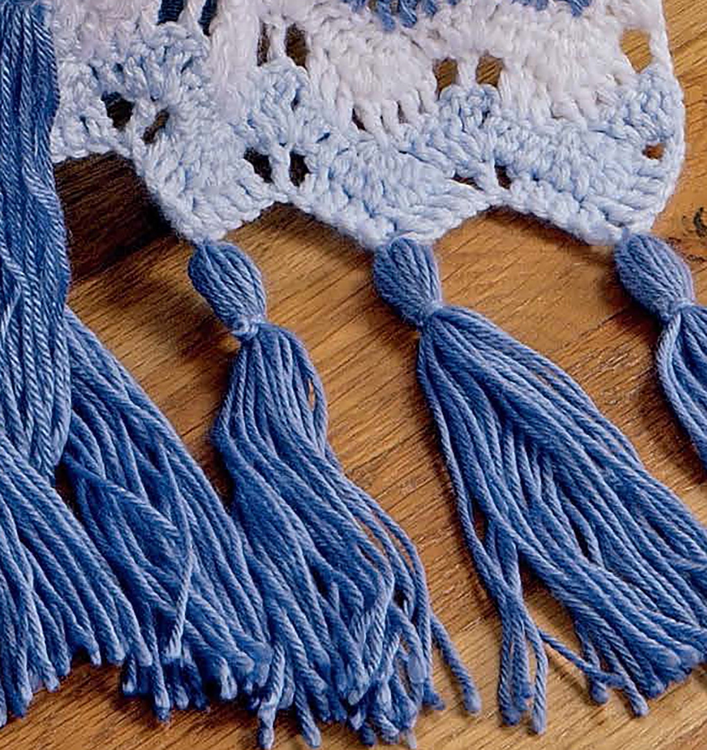 Displayed on a wooden surface, the "Learn to Crochet Ripple Afghans" digital download by Leisure Arts features blue and white crocheted fabric with long blue tassels—perfect inspiration for your next ripple afghan project.