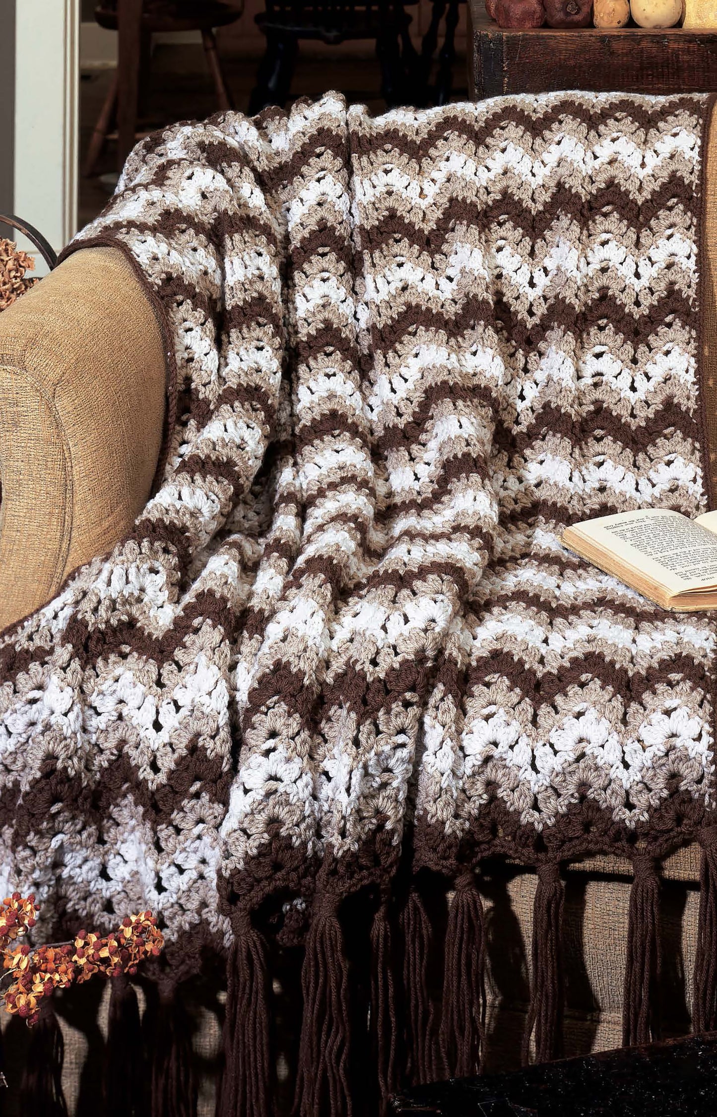 The tan sofa is adorned with a brown and white zigzag blanket, made using the "Learn to Crochet Ripple Afghans" digital pattern by Leisure Arts. An open book rests nearby, adding to the cozy and inviting vibe.